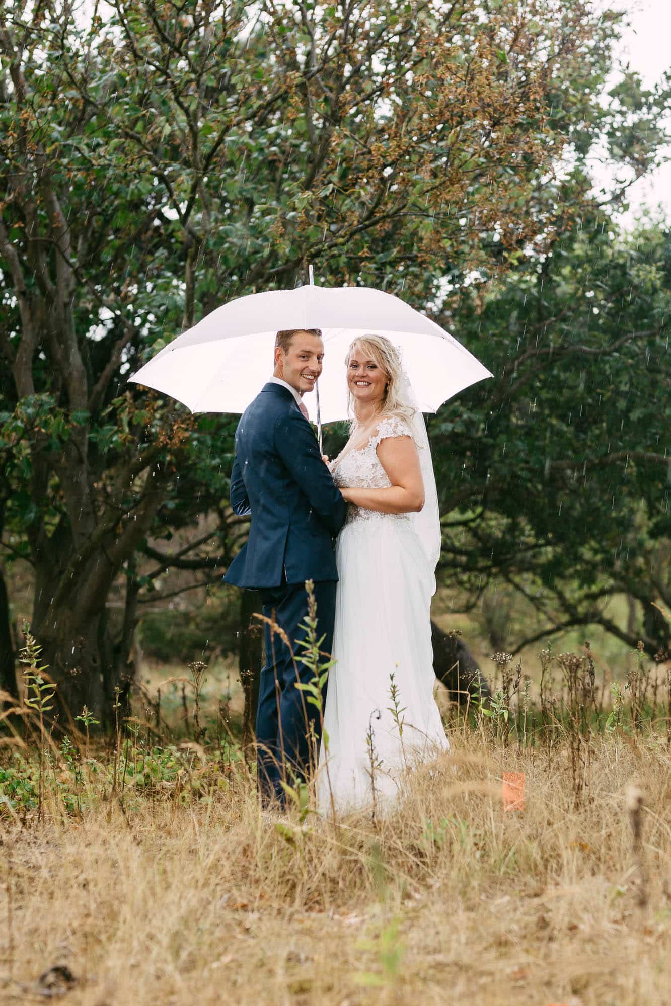 A bride and groom stand under an umbrella in a field.