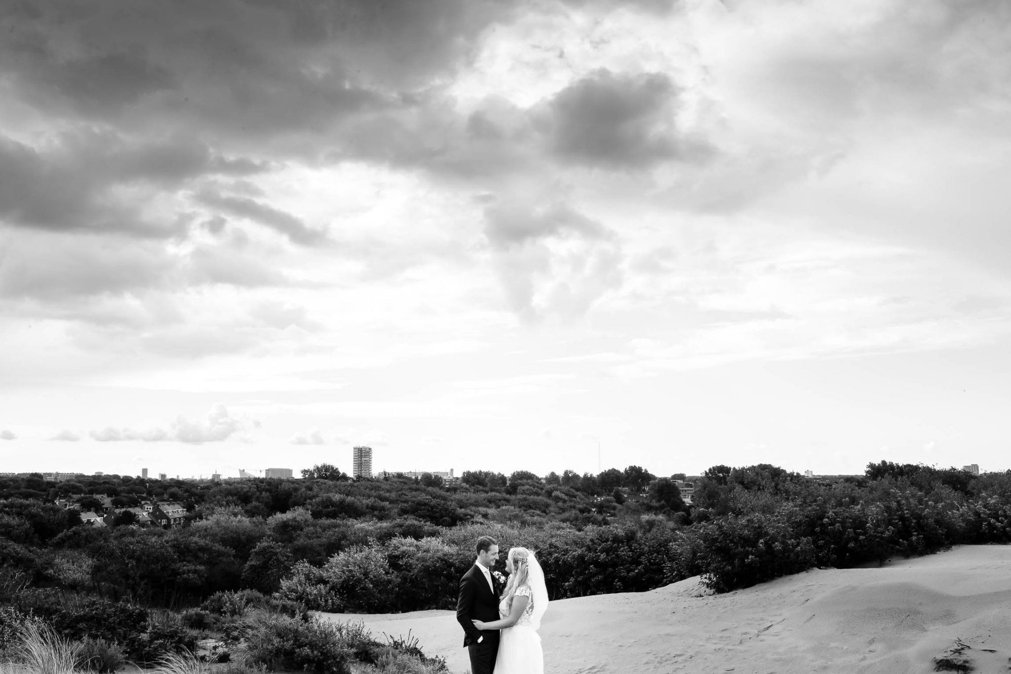A bride and groom stand on a sand dune under a cloudy sky.