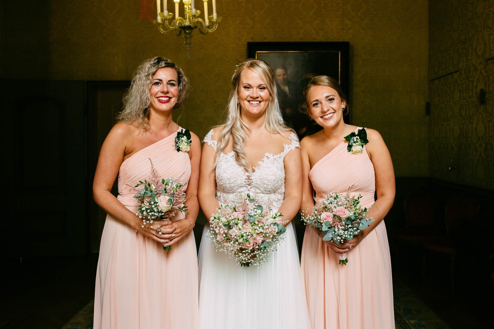 Three bridesmaids in pink dresses stand in an ornate room.