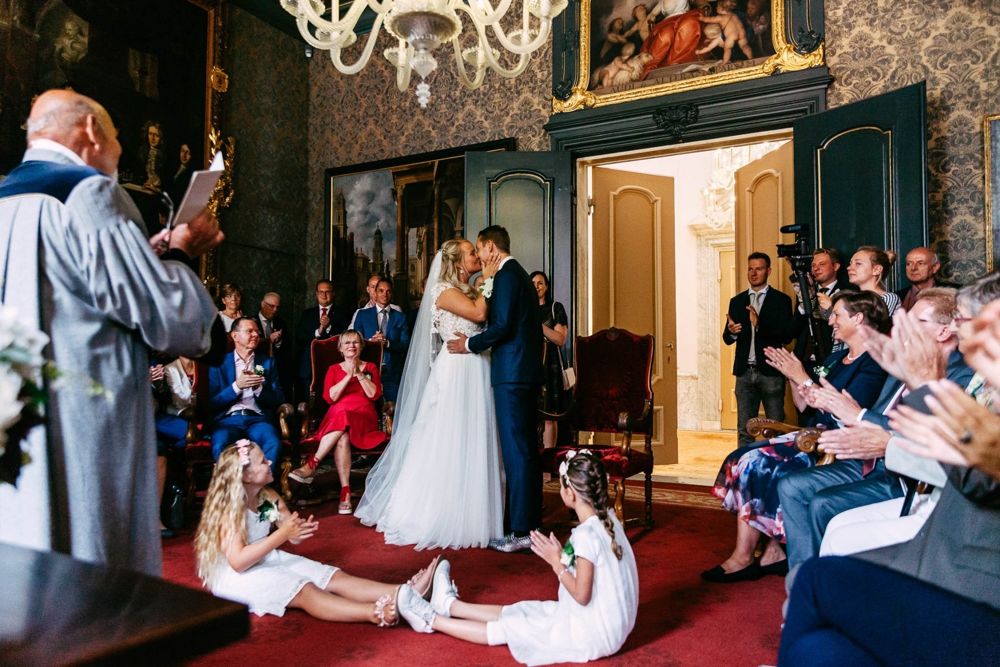 A wedding ceremony in an ornate room with a kissing bride and groom.