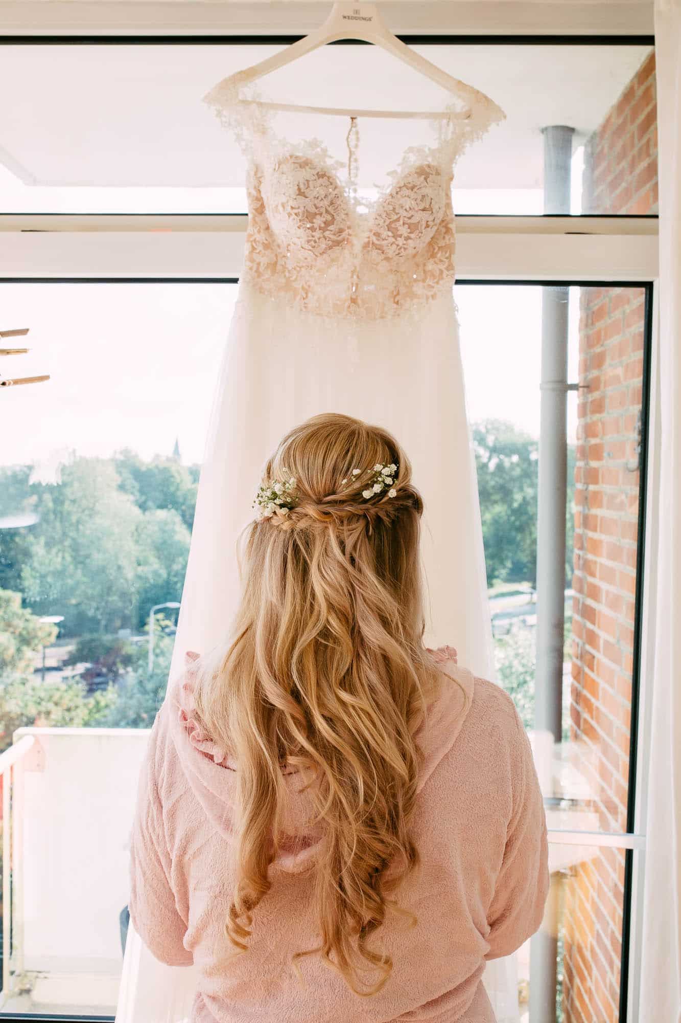 A bride looks at her wedding dress in front of a window.
