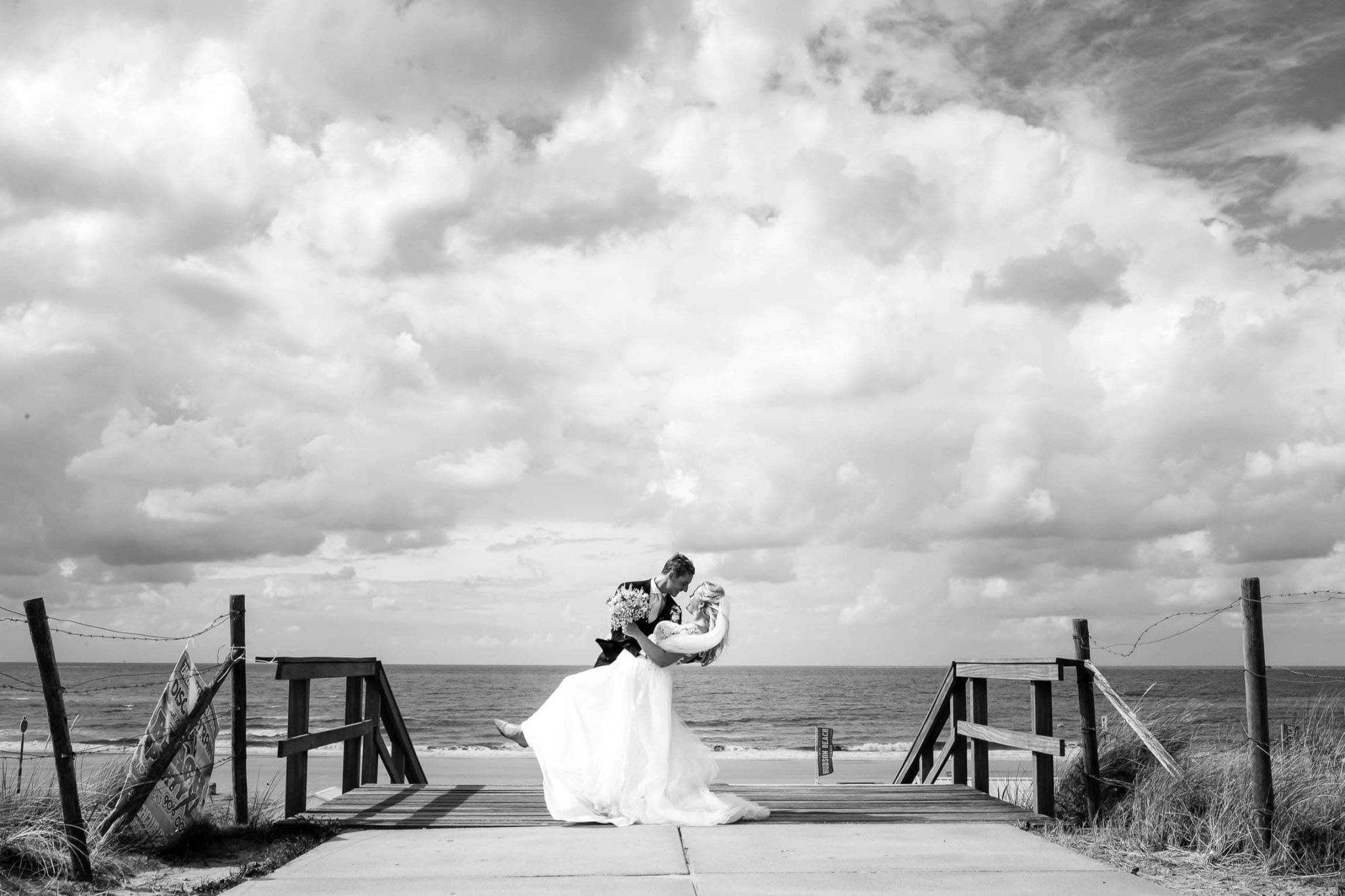 A bride and groom kiss on a boardwalk near the ocean.
