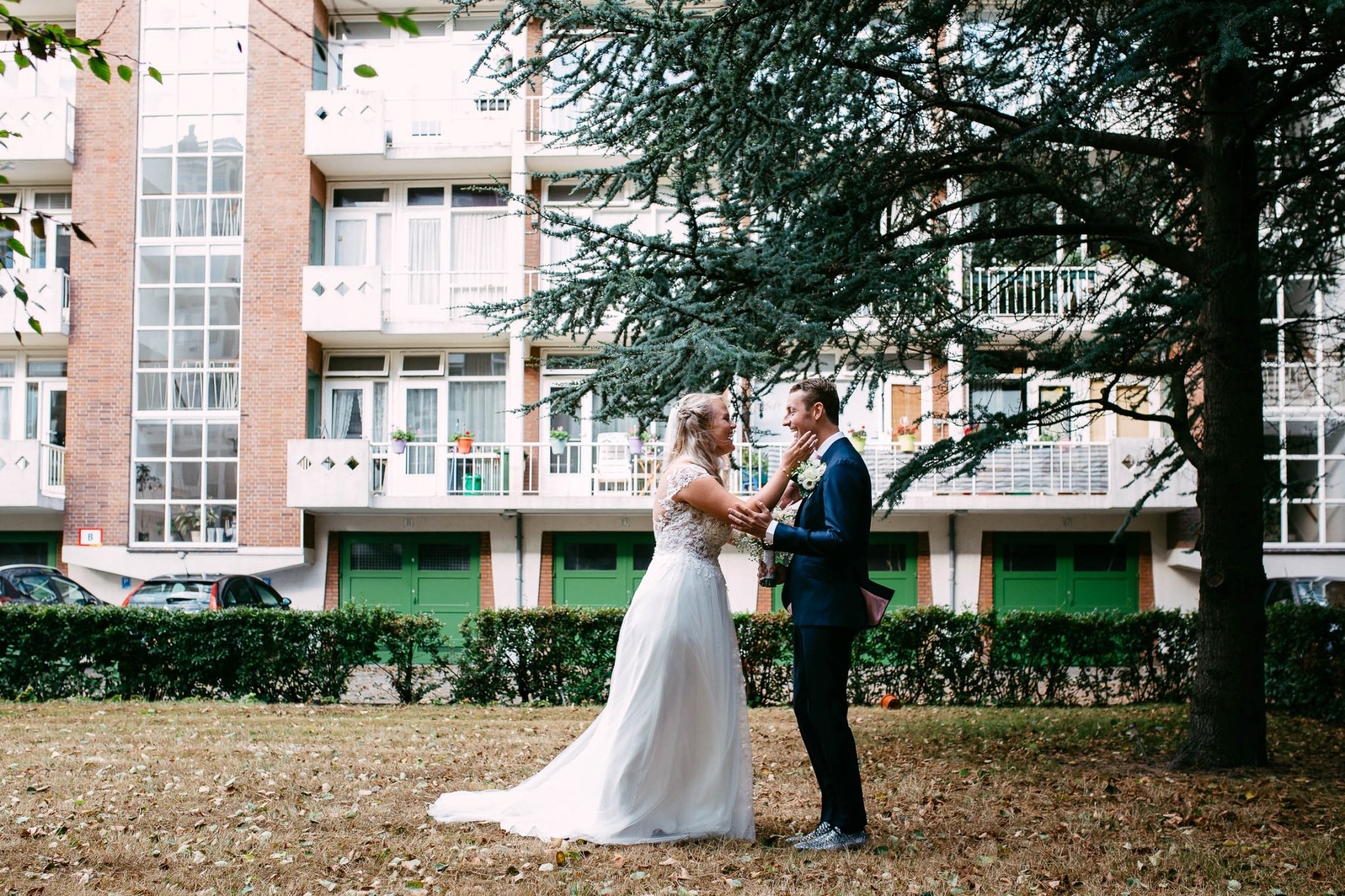 A bride and groom stand in front of an apartment complex.