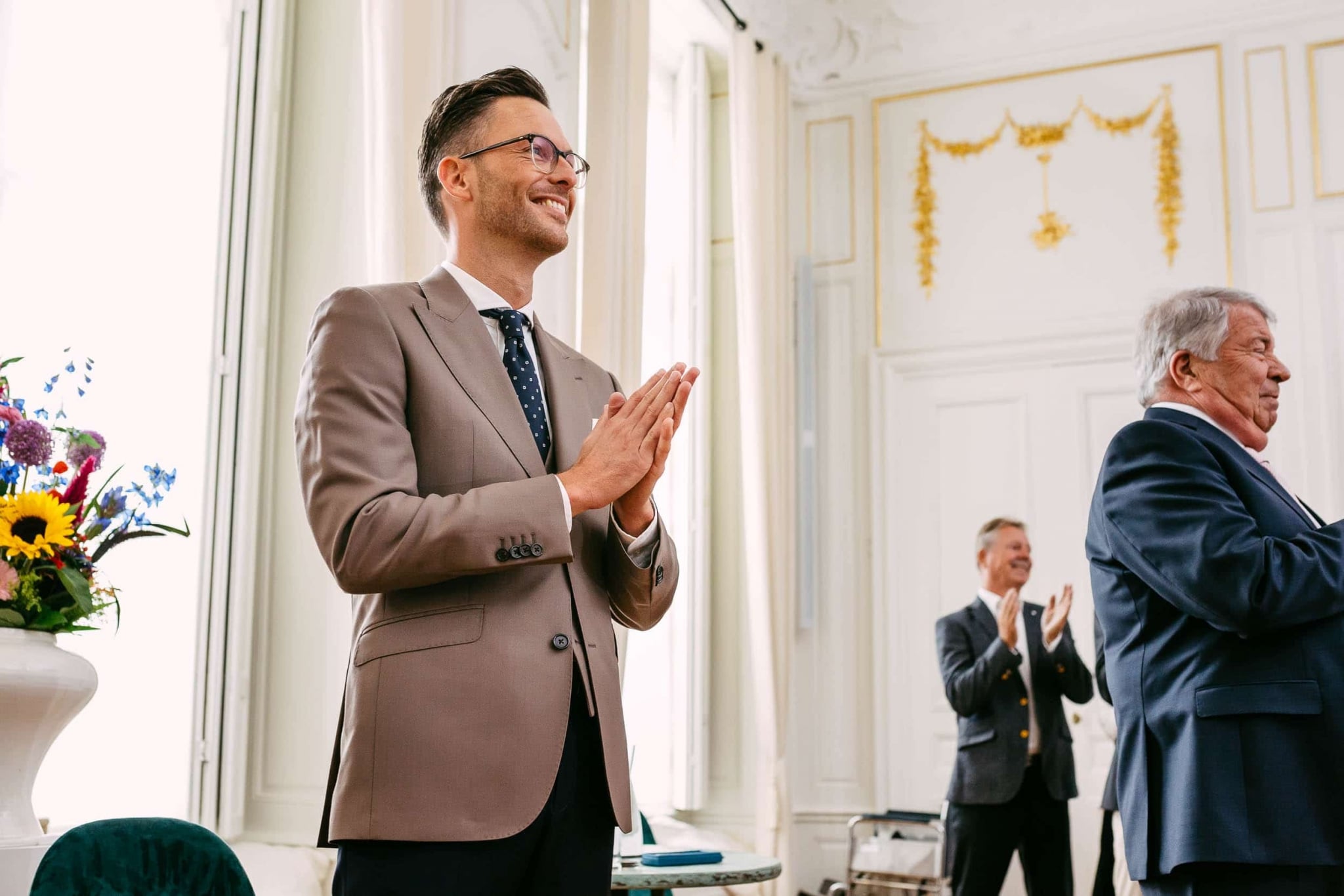 A man in a suit applauds another man in a room.