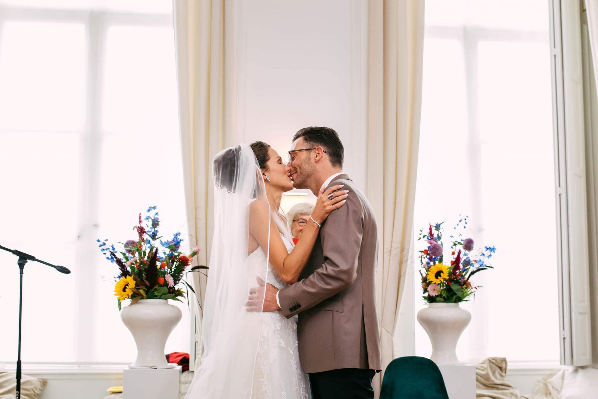 A bride and groom kiss in front of large windows.