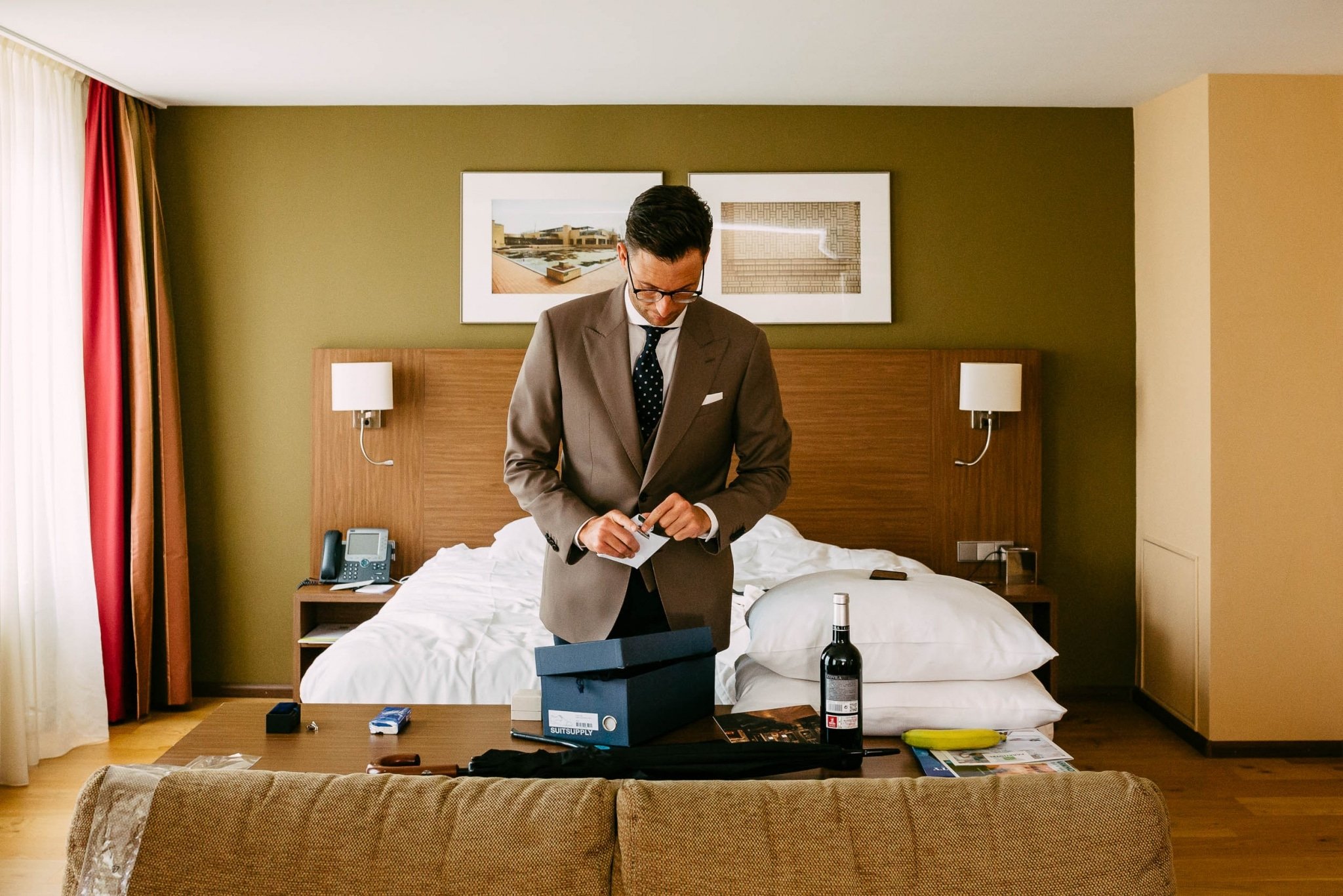 Een man in een pak staat naast een bed in een hotelkamer.