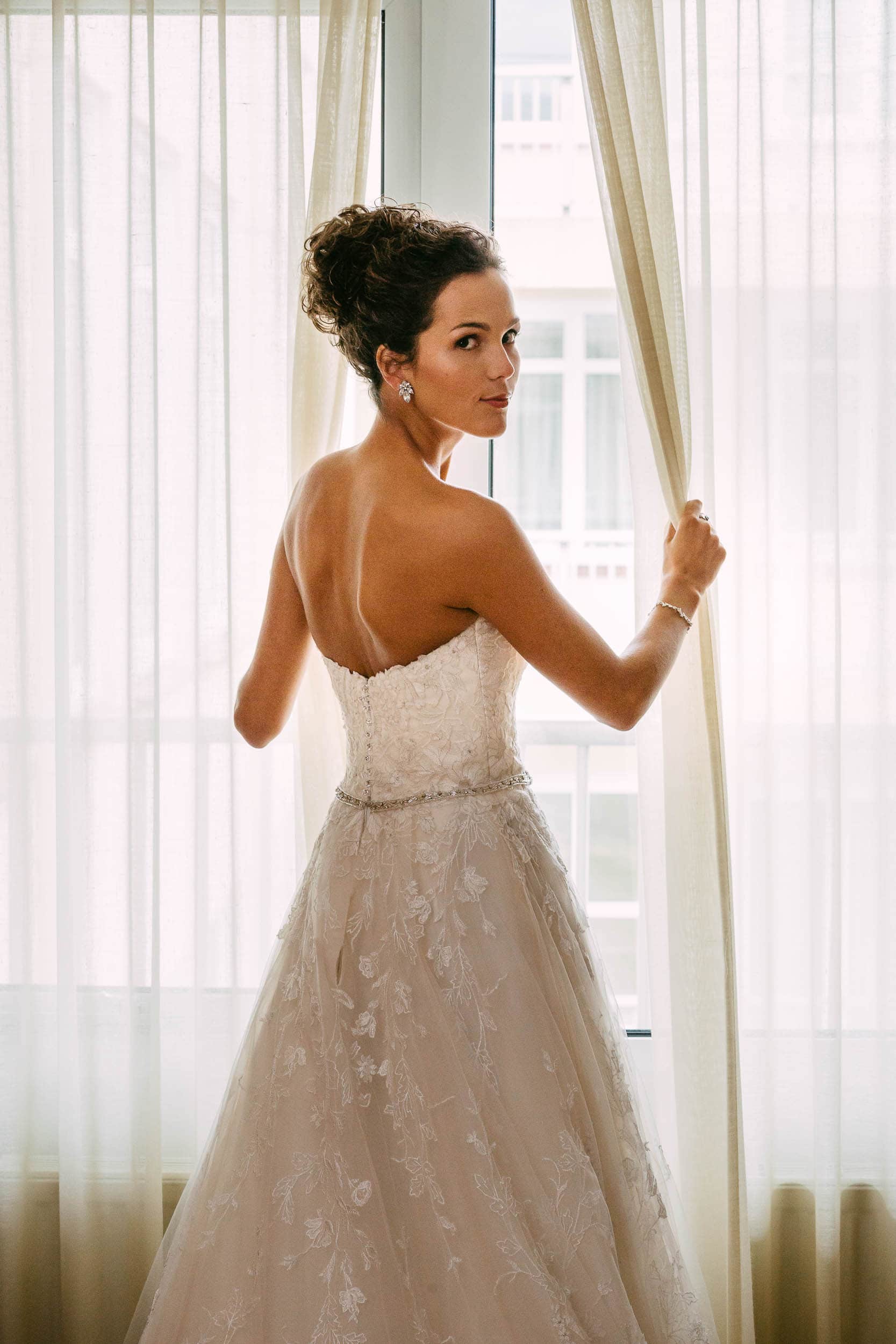 A bride in a wedding dress stands in front of a window.