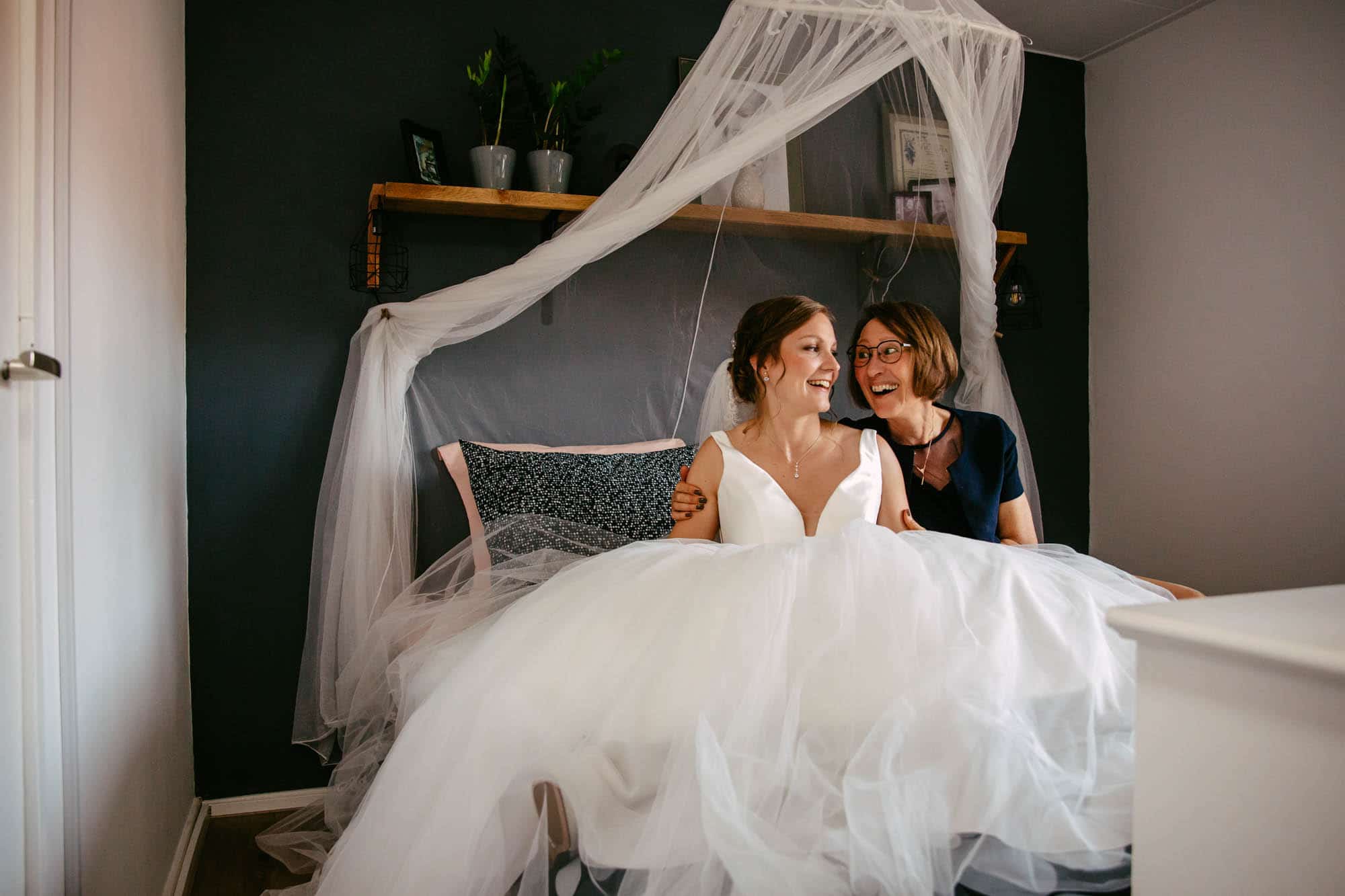 A bride and her mother sit on a bed in a bedroom.