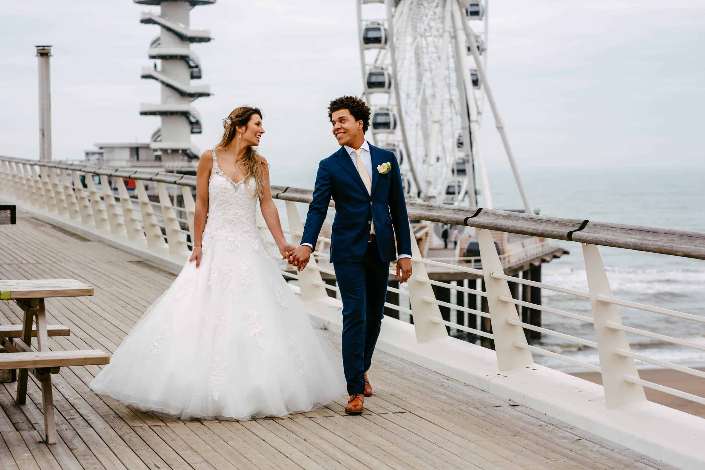 A bride and groom walk on a boardwalk near a Ferris wheel.