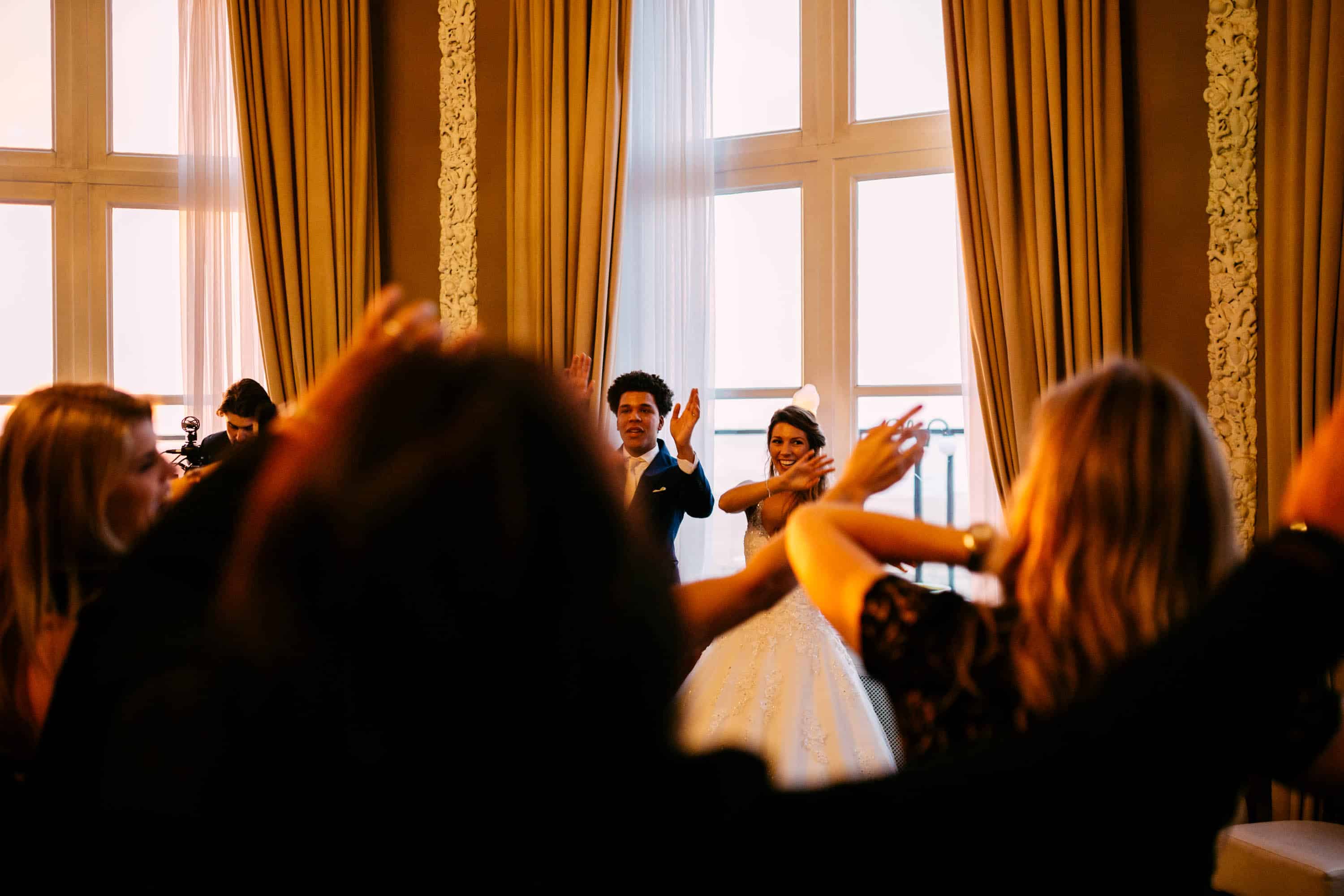 Een bruid en bruidegom zwaaien met hun handen in de lucht tijdens een huwelijksreceptie.