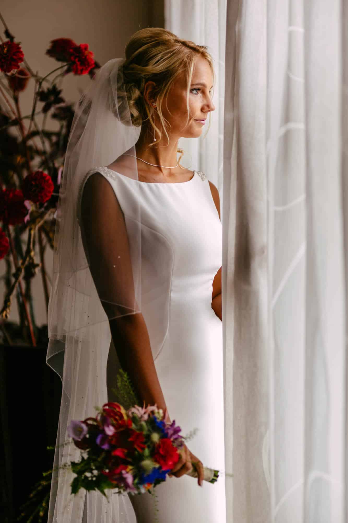 A bride in a white dress stands in front of a window.