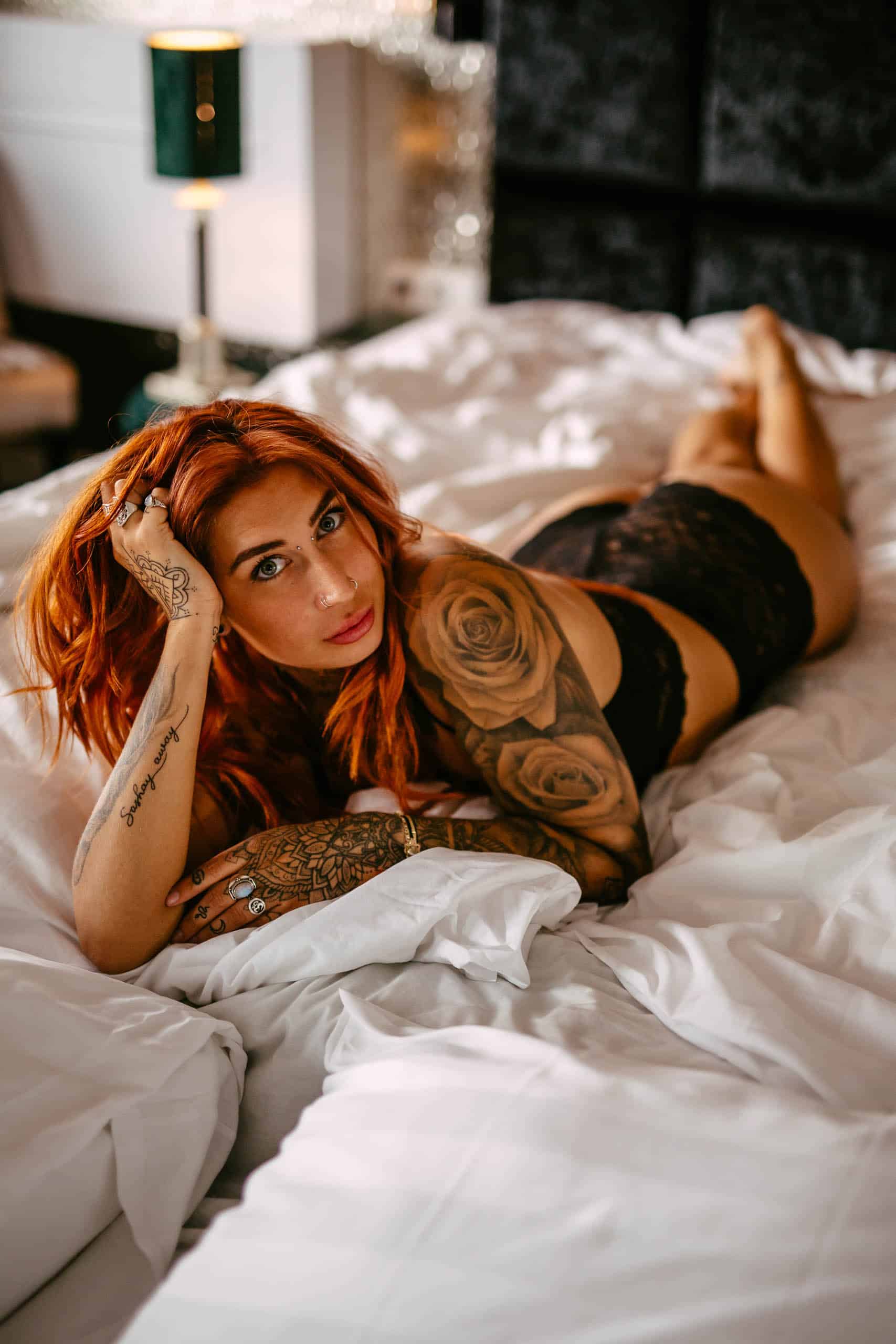 A woman with red hair and tattoos poses sensually on a bed during a captivating boudoir shoot in Hoek van Holland.
