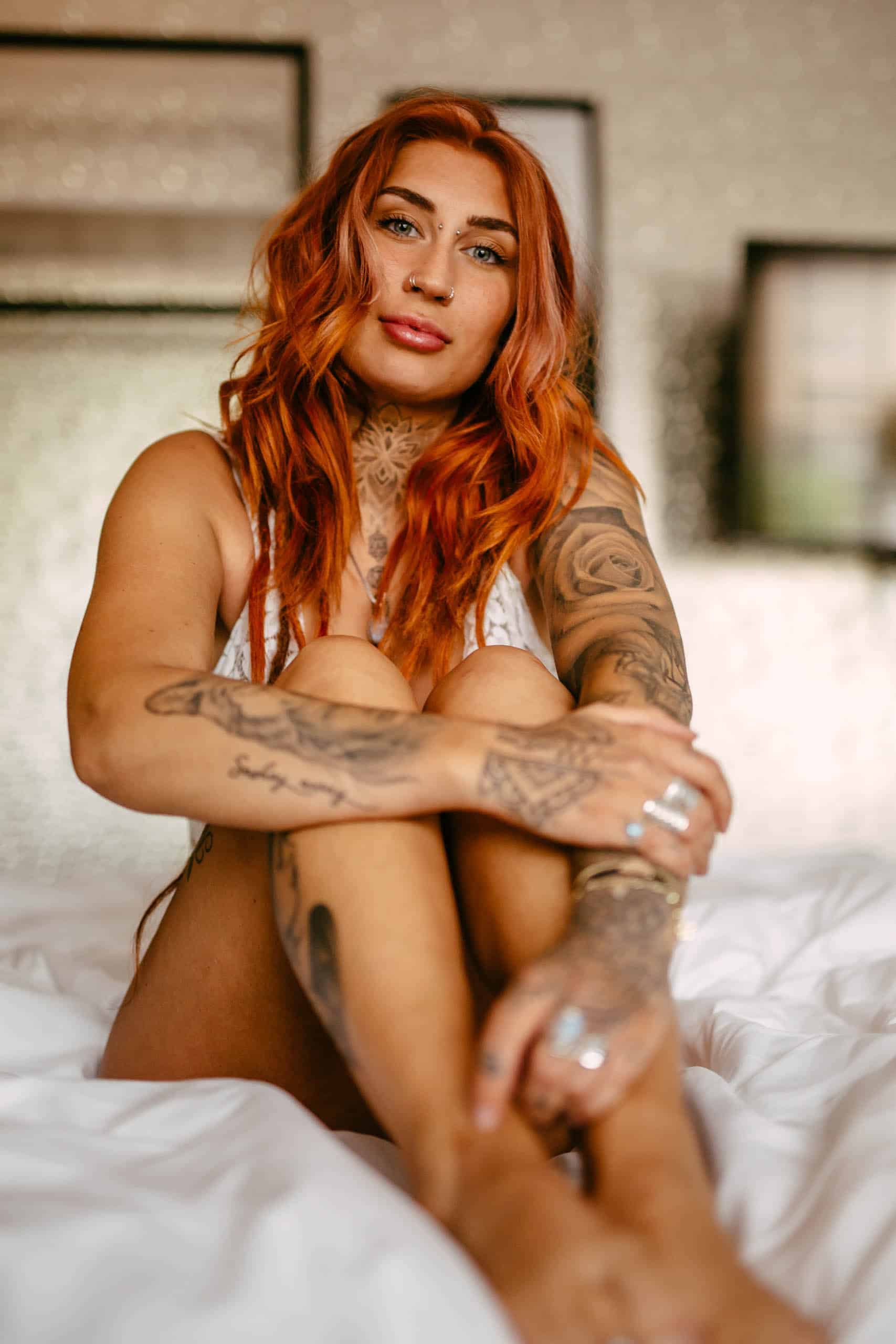 A woman with red hair sitting on a bed during a boudoir shoot in Hoek van Holland, showing off her beautiful tattoos.