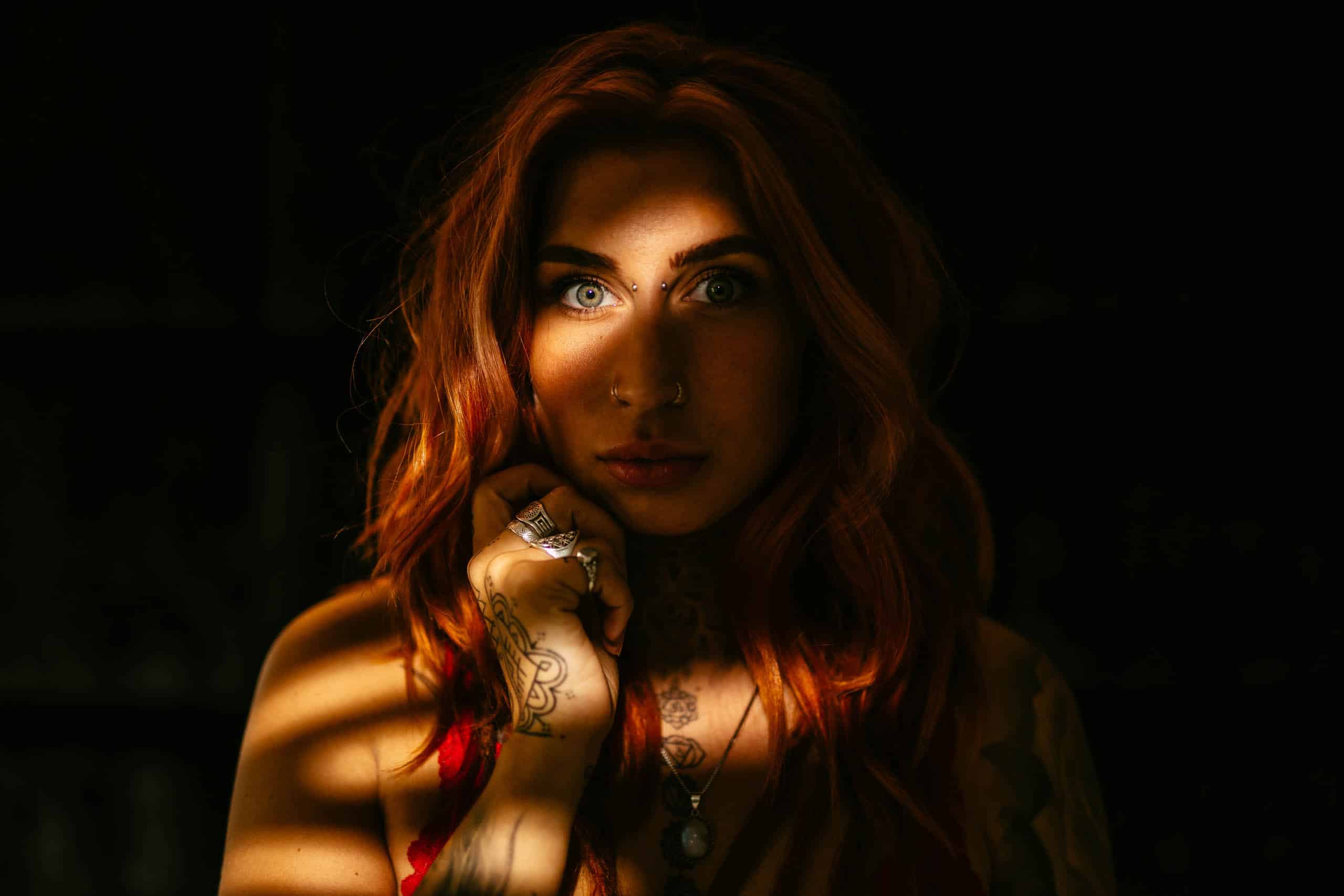 A woman with red hair and tattoos poses in a dark room during a boudoir shoot in Hoek van Holland.