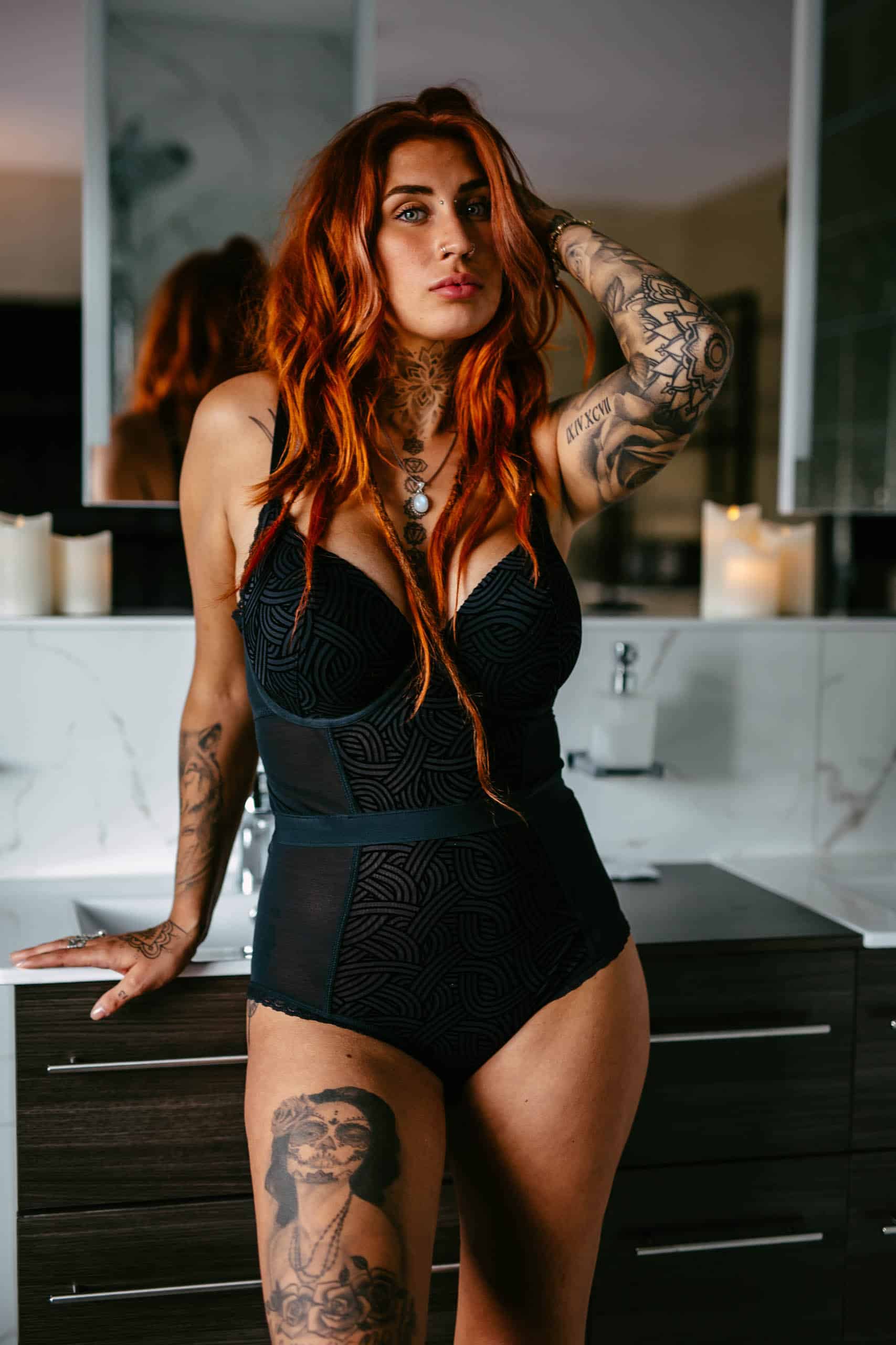 A woman with red hair and tattoos poses in a bathroom during a boudoir shoot.