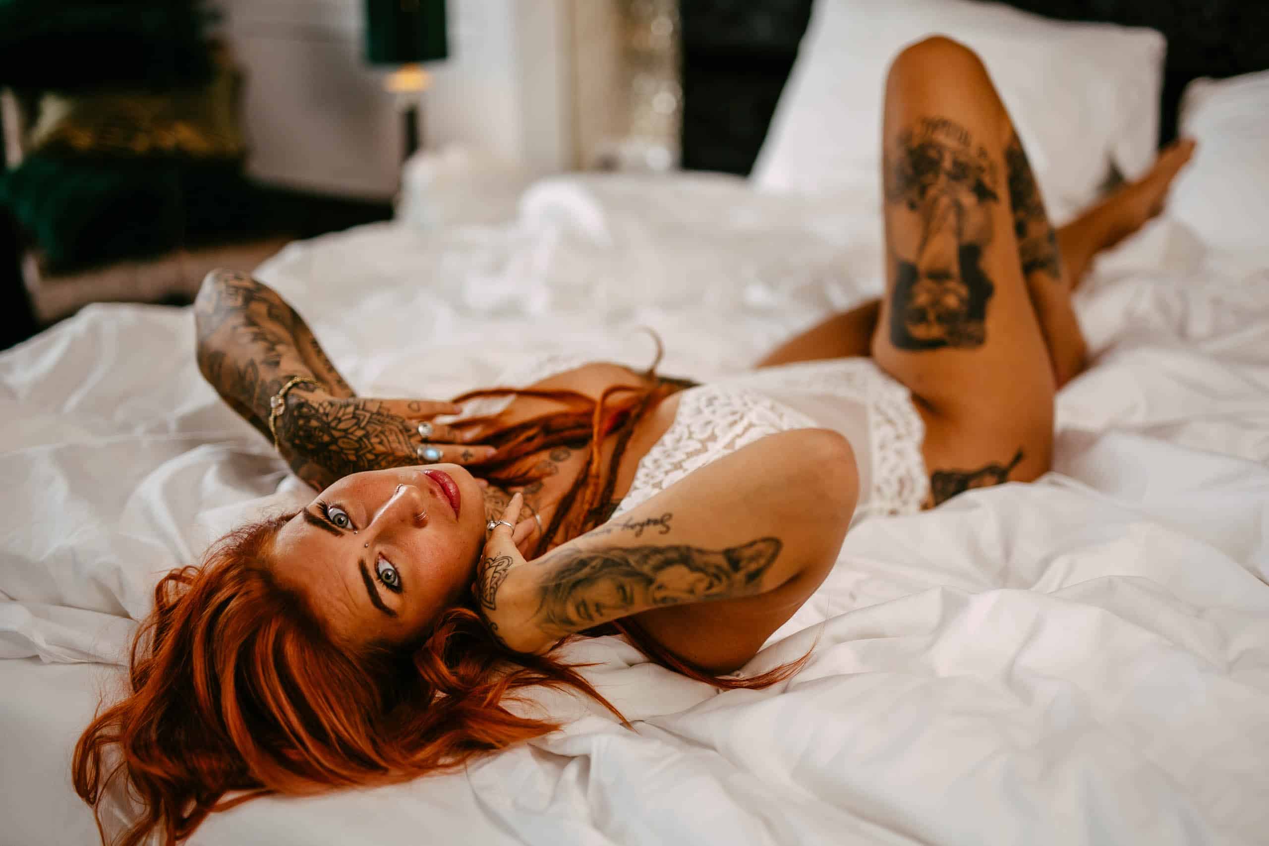 A woman with red hair and tattoos doing a boudoir shoot in Hoek van Holland while lying on her bed.