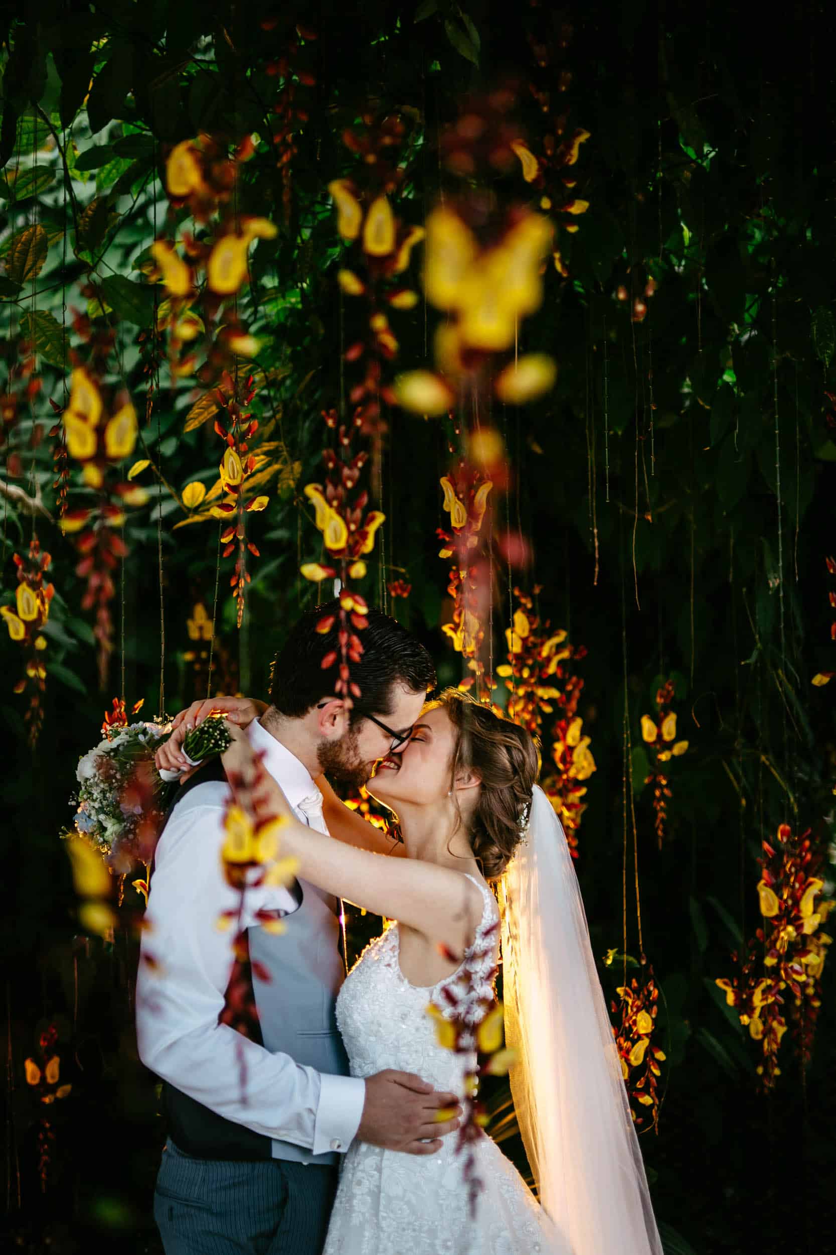 Een bruid en bruidegom kussen elkaar onder een bladerdak van bloemen bij een Trouwerij in de botanische tuin delft.