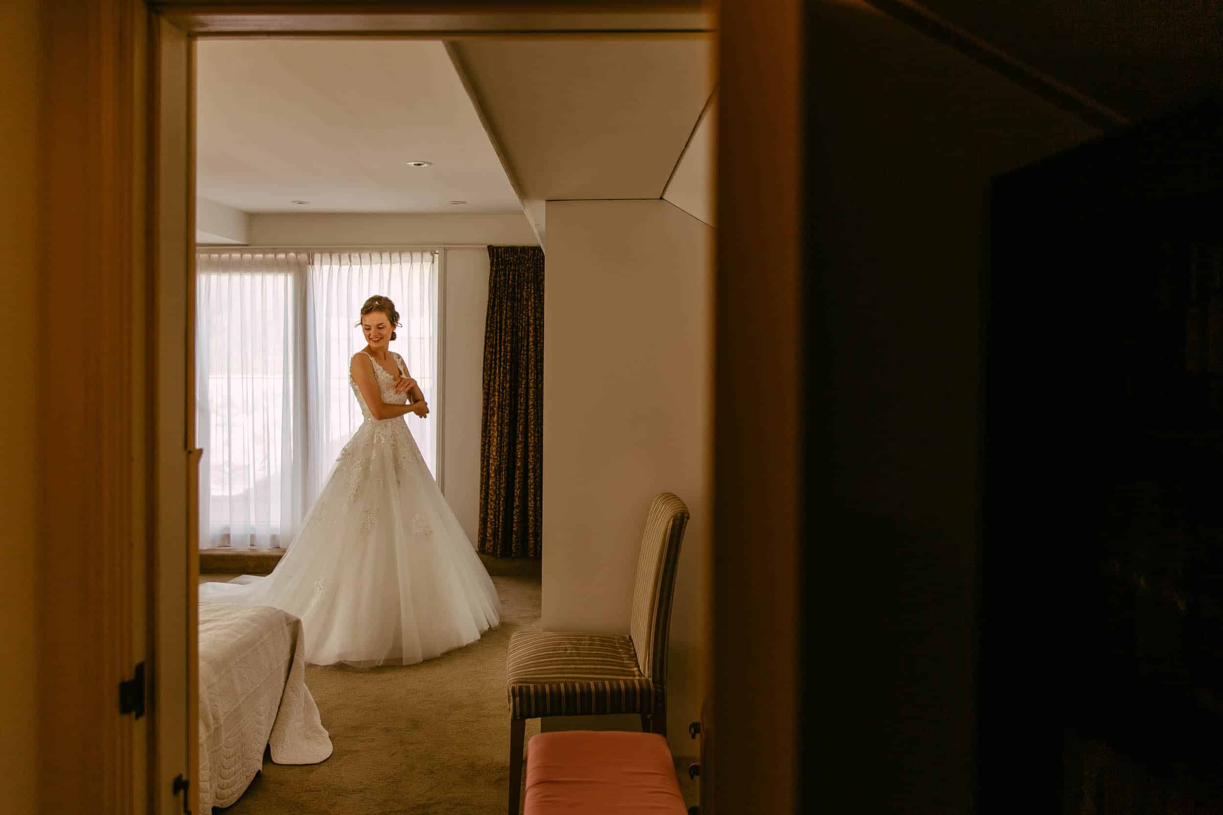 A bride in wedding dress stands in the doorway of a hotel room, ready for her wedding at the botanical garden delft.