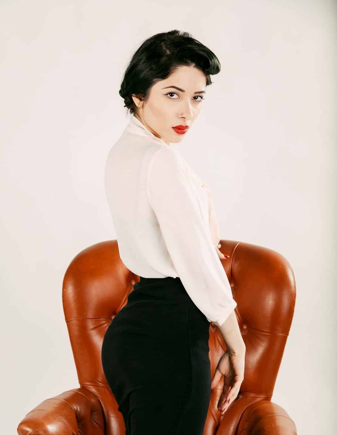 A professional portrait photographer captures the image of a woman in a black skirt and white shirt as she poses conspicuously on a chair.