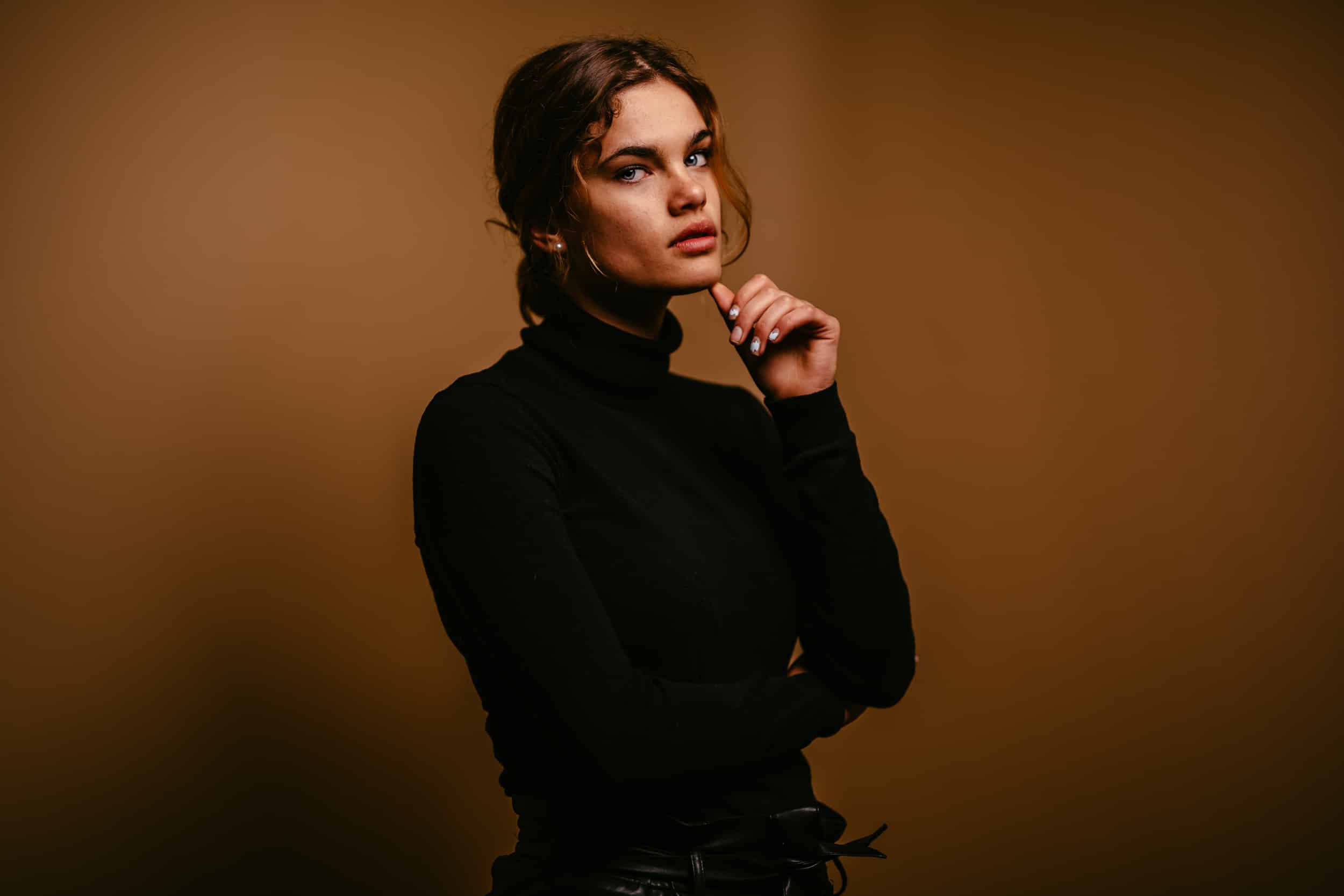Een jonge vrouw in een zwarte coltrui poseert professionele portretfotograaf met haar hand op haar kin.