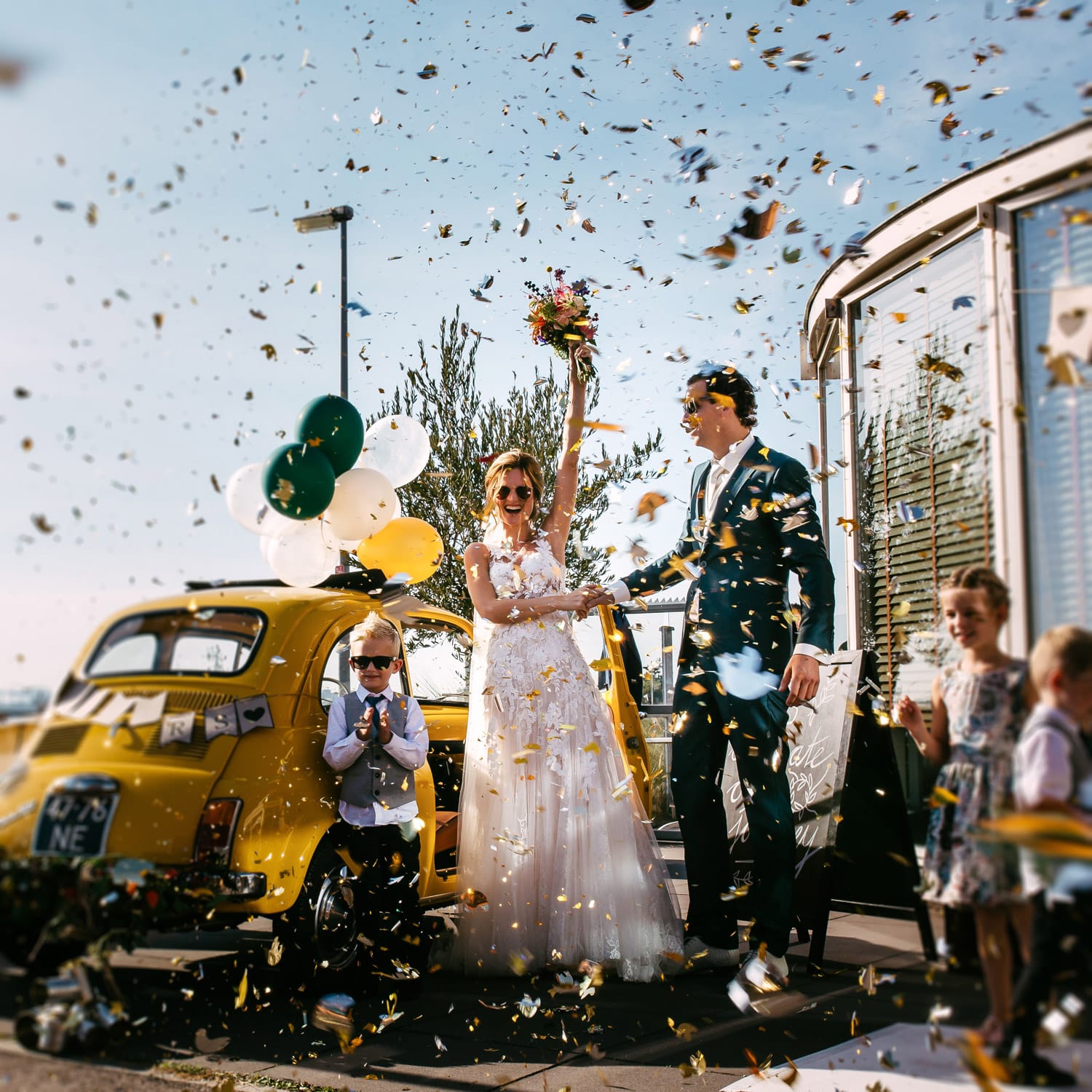 Een bruid en bruidegom staan voor een gele auto, vastgelegd door een professionele trouwfotograaf terwijl er vrolijk confetti naar hen wordt gegooid.