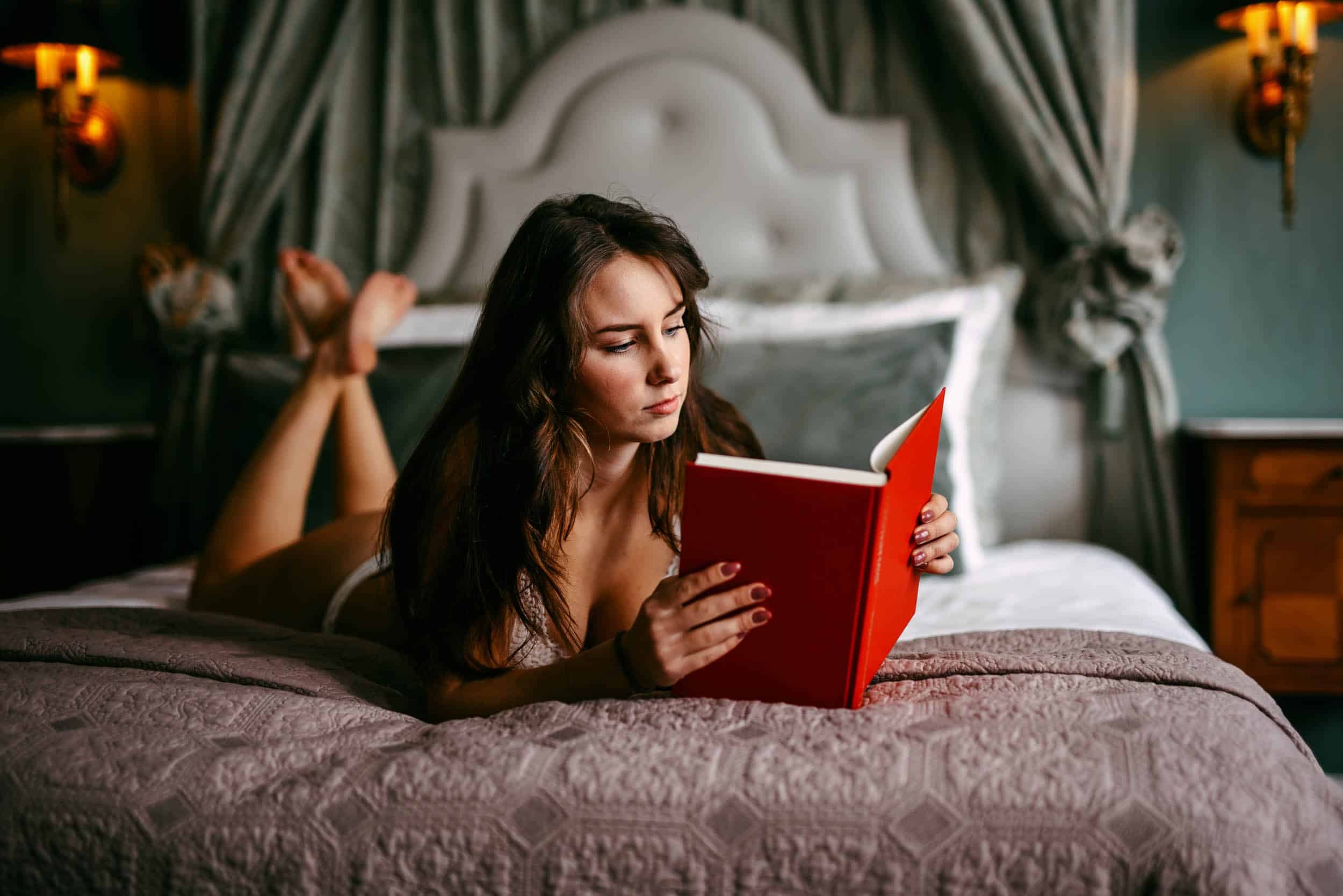 A woman lying on bed reading a book in a boudoir.