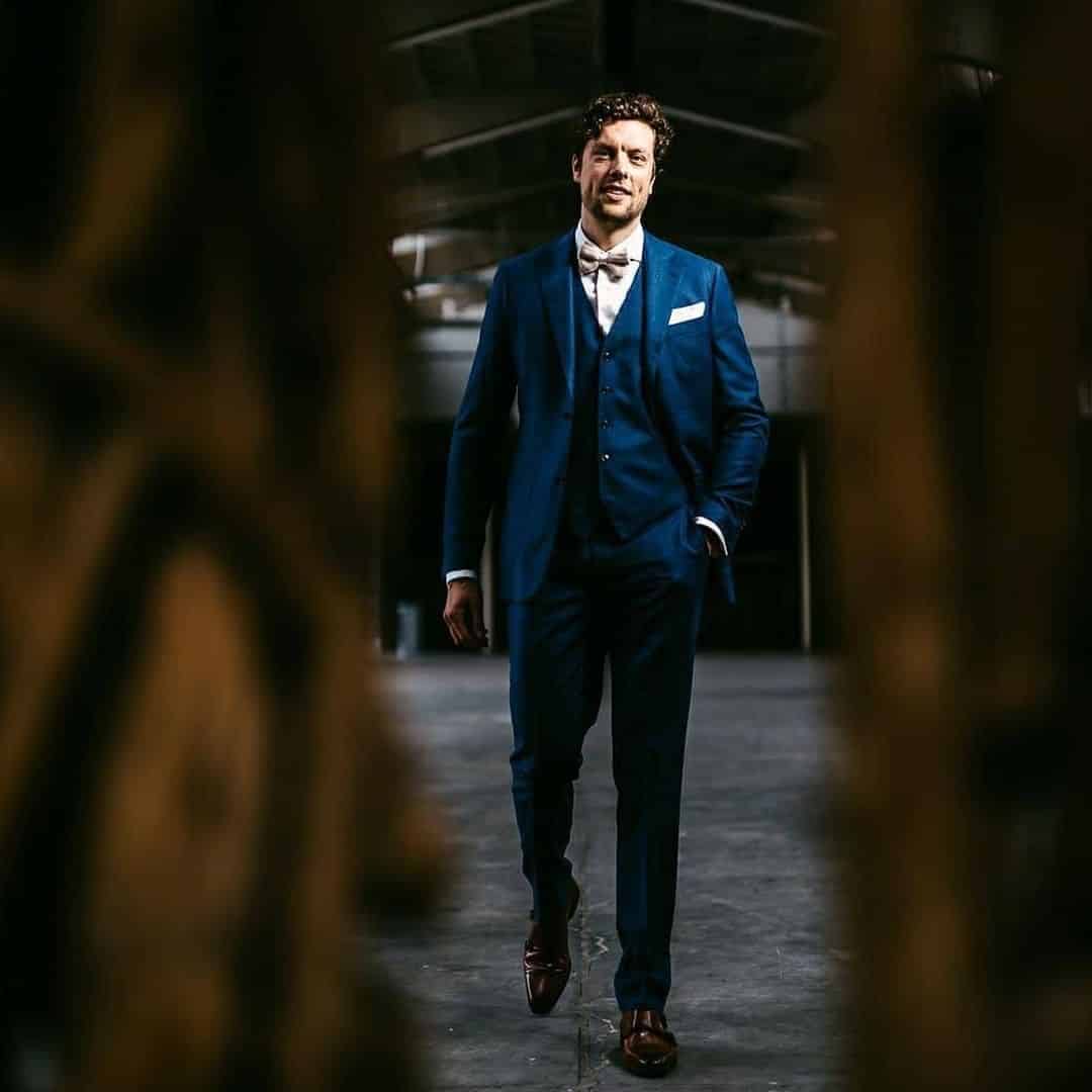 A man, dressed in a blue suit, poses confidently in a warehouse for an Instagram-worthy photo shoot captured by a talented wedding photographer.