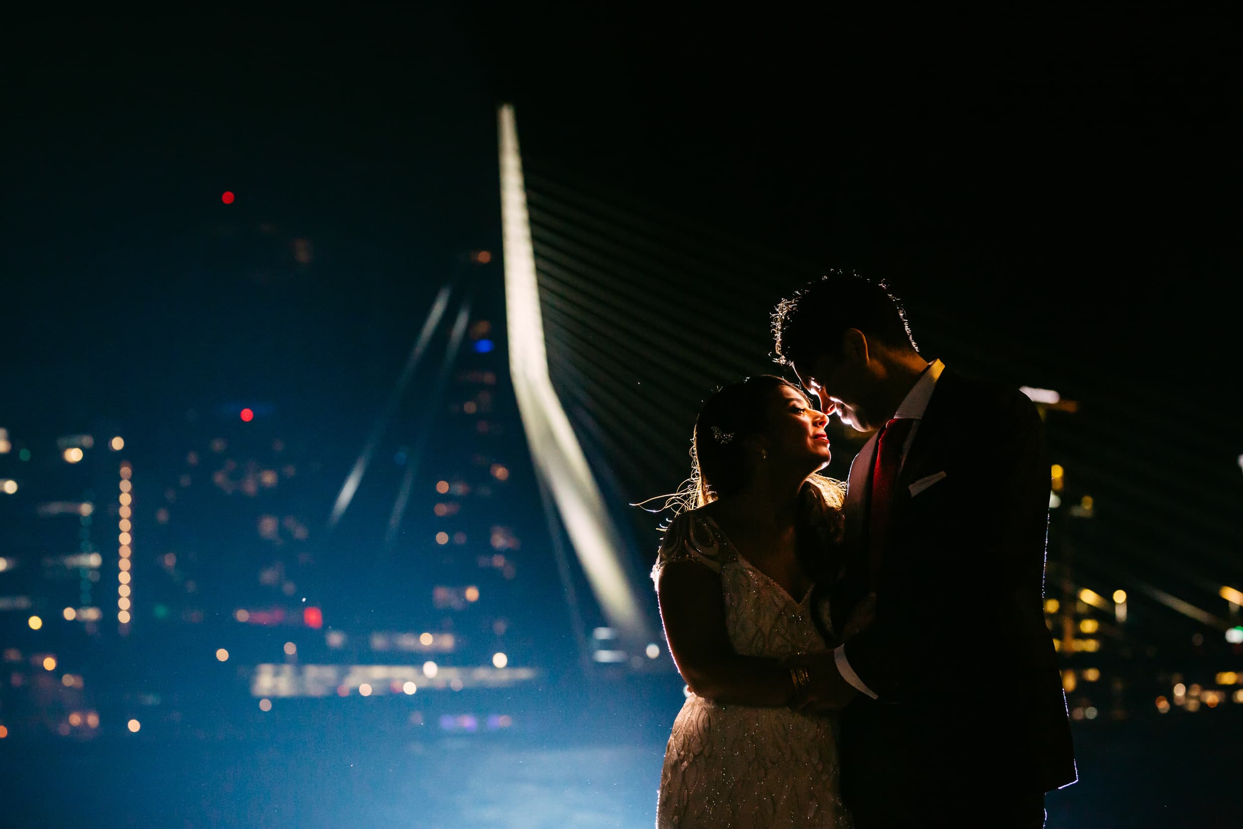 A bride and groom stand in front of a bridge in Rotterdam at night.