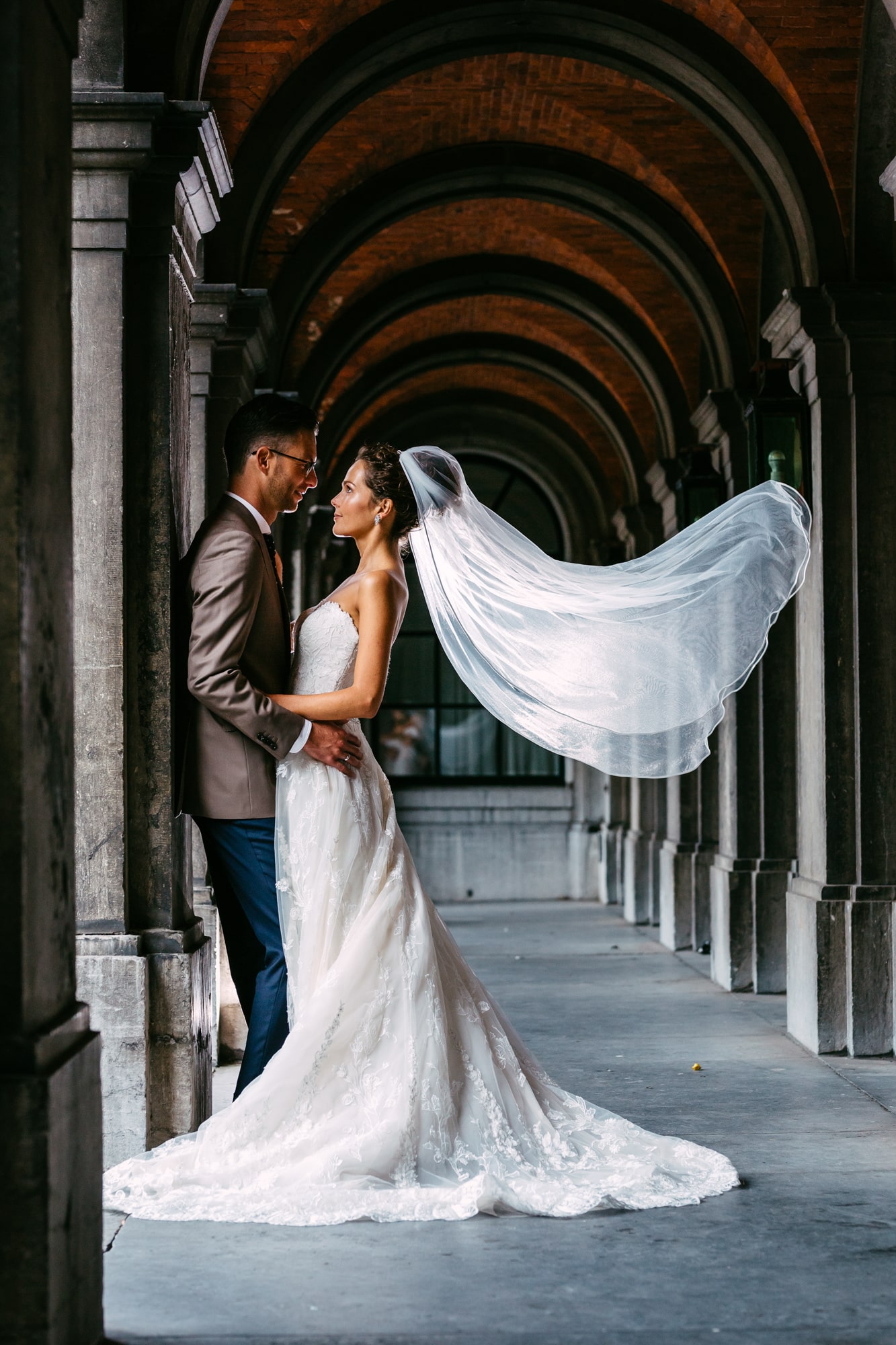 Een bruid en bruidegom, vastgelegd in prachtige trouwfotografie, poseren onder de bogen terwijl hun sluier sierlijk opwaait in de wind.