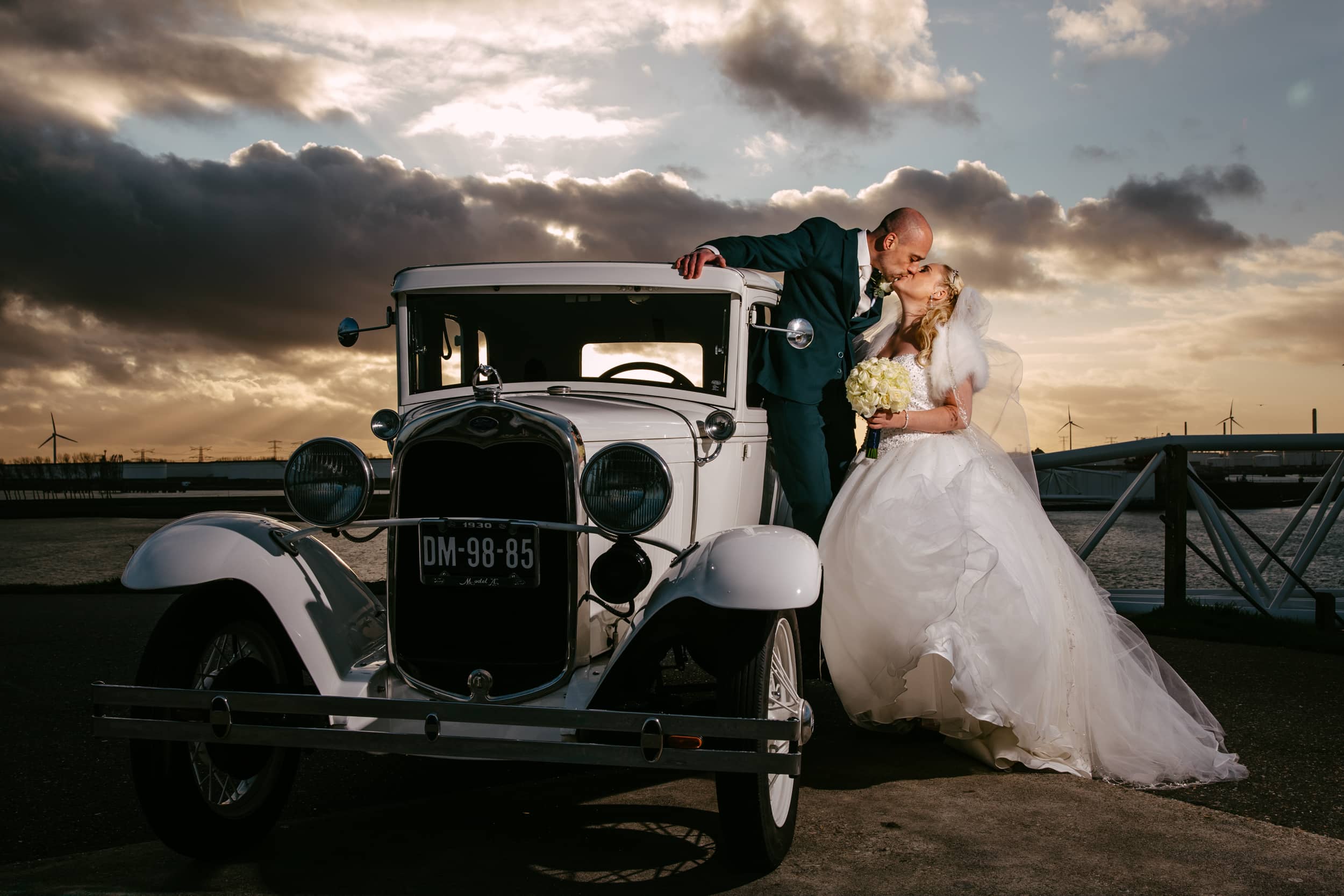 A bride and groom pose in contact with a vintage car and share a romantic kiss.