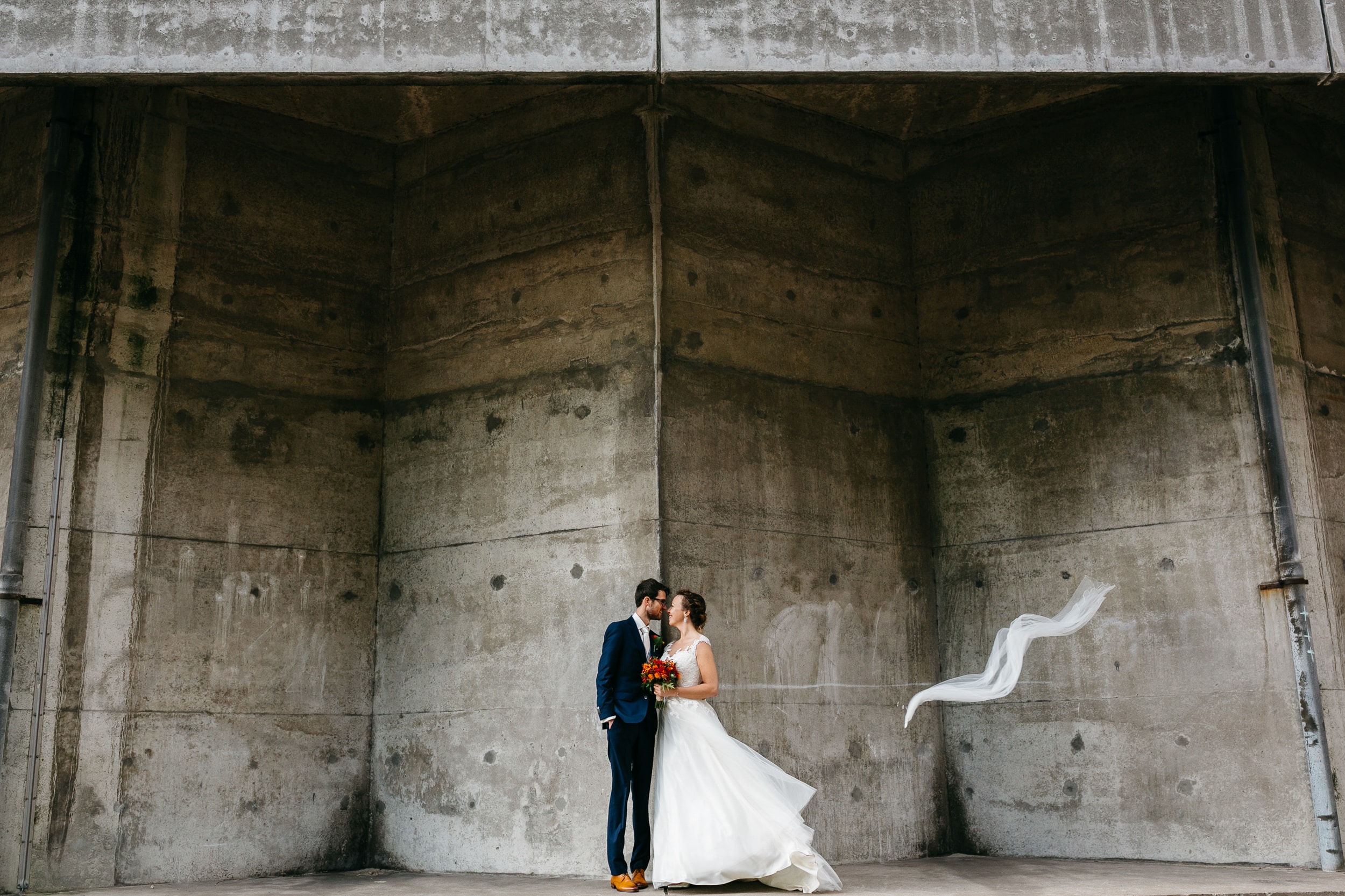 A bride and groom pose elegantly in front of a concrete wall, with the bride wearing a beautiful A-line wedding dress.