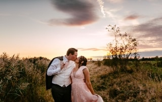 Trouwfotos: Een bruid en bruidegom die elkaar hartstochtelijk kussen in een veld bij zonsondergang.