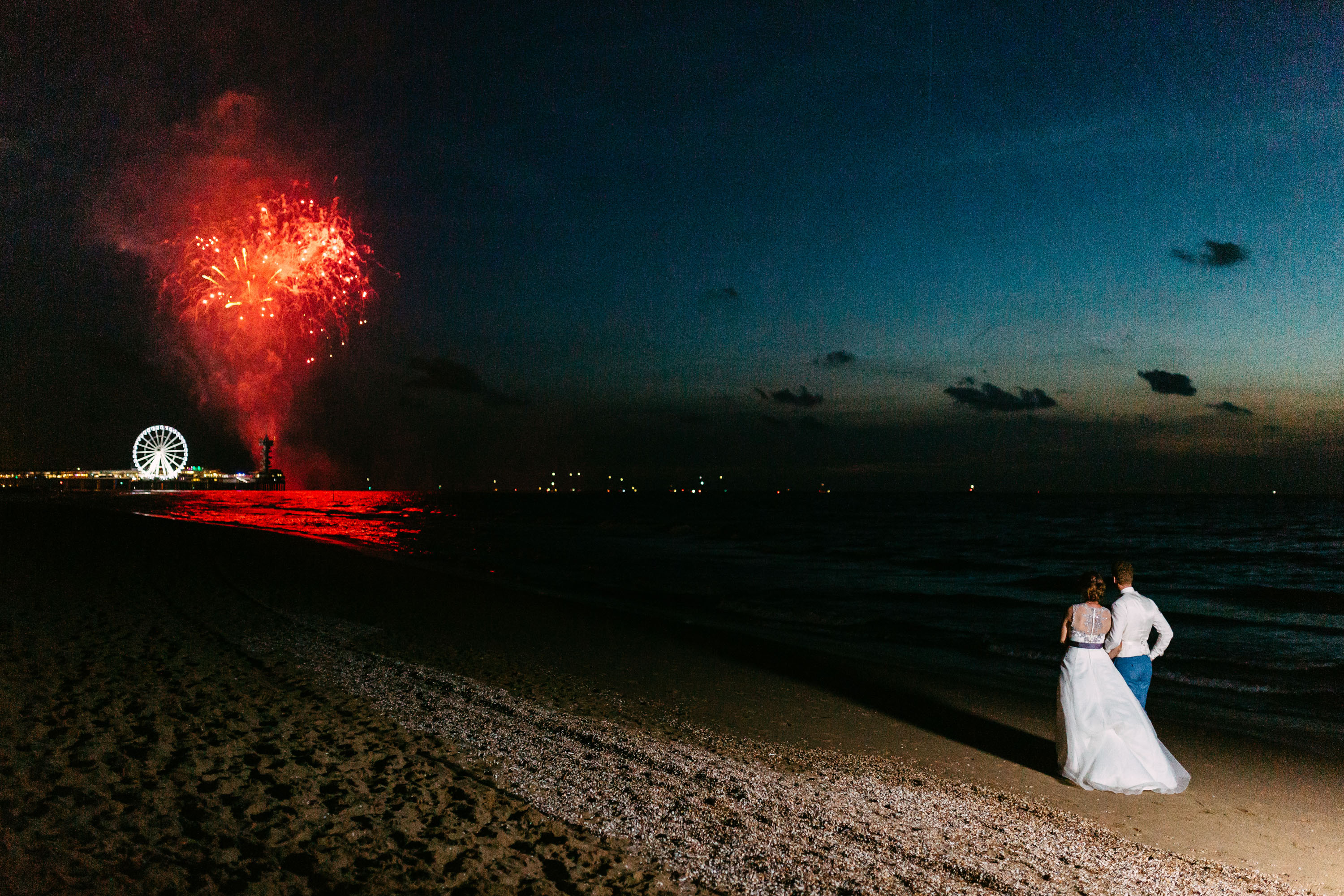 A bride and groom stand on the beach during a wedding shoot, with fireworks in the background.