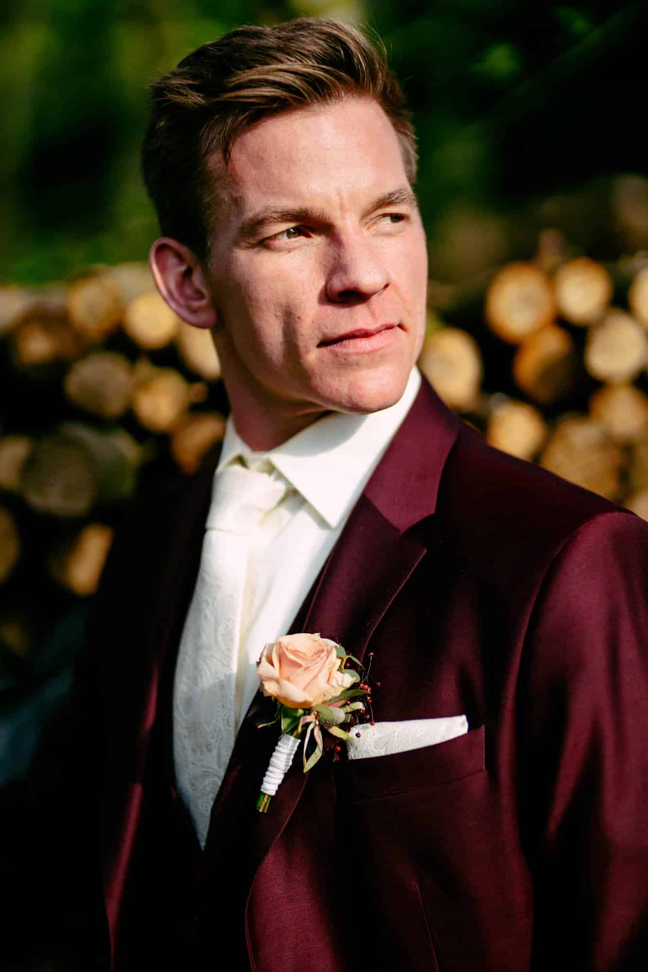 A man in a burgundy suit stands in front of tree trunks.