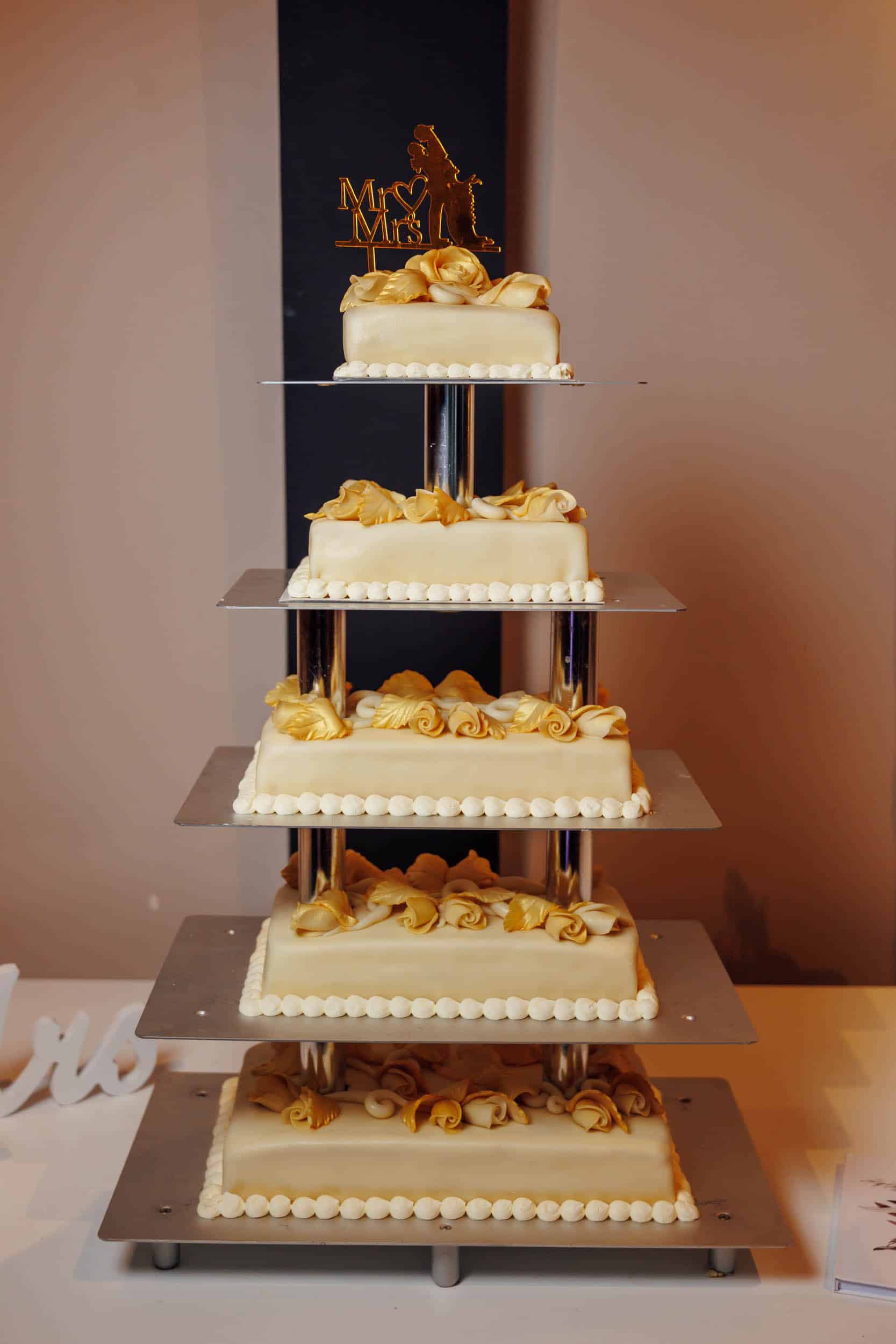 A five-tier wedding cake decorated with gold roses and pearls, displayed on a metal stand with "mr & mrs" sign on the background.