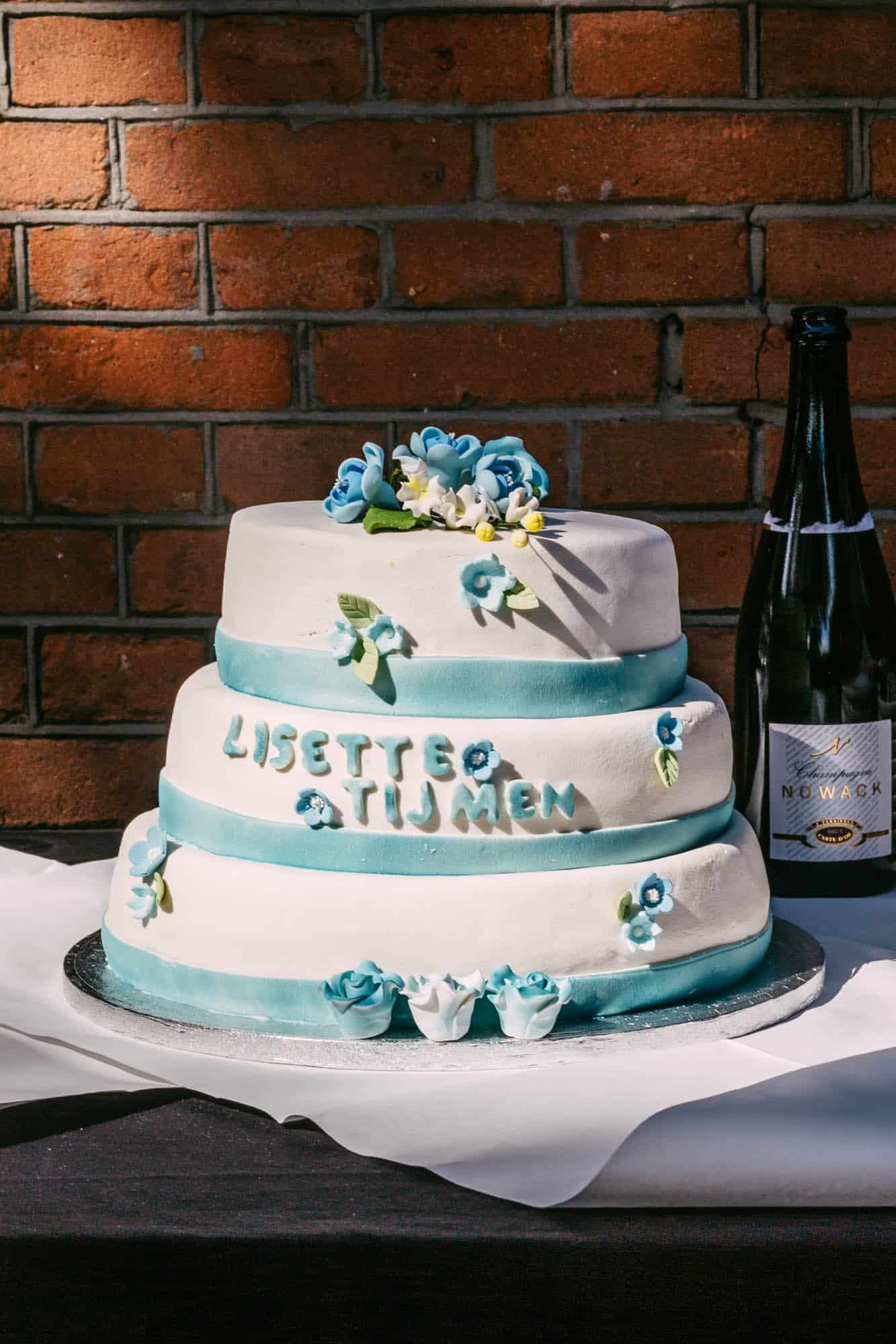 Beschrijving: Een bruidstaart met blauwe bloemen en een fles champagne.