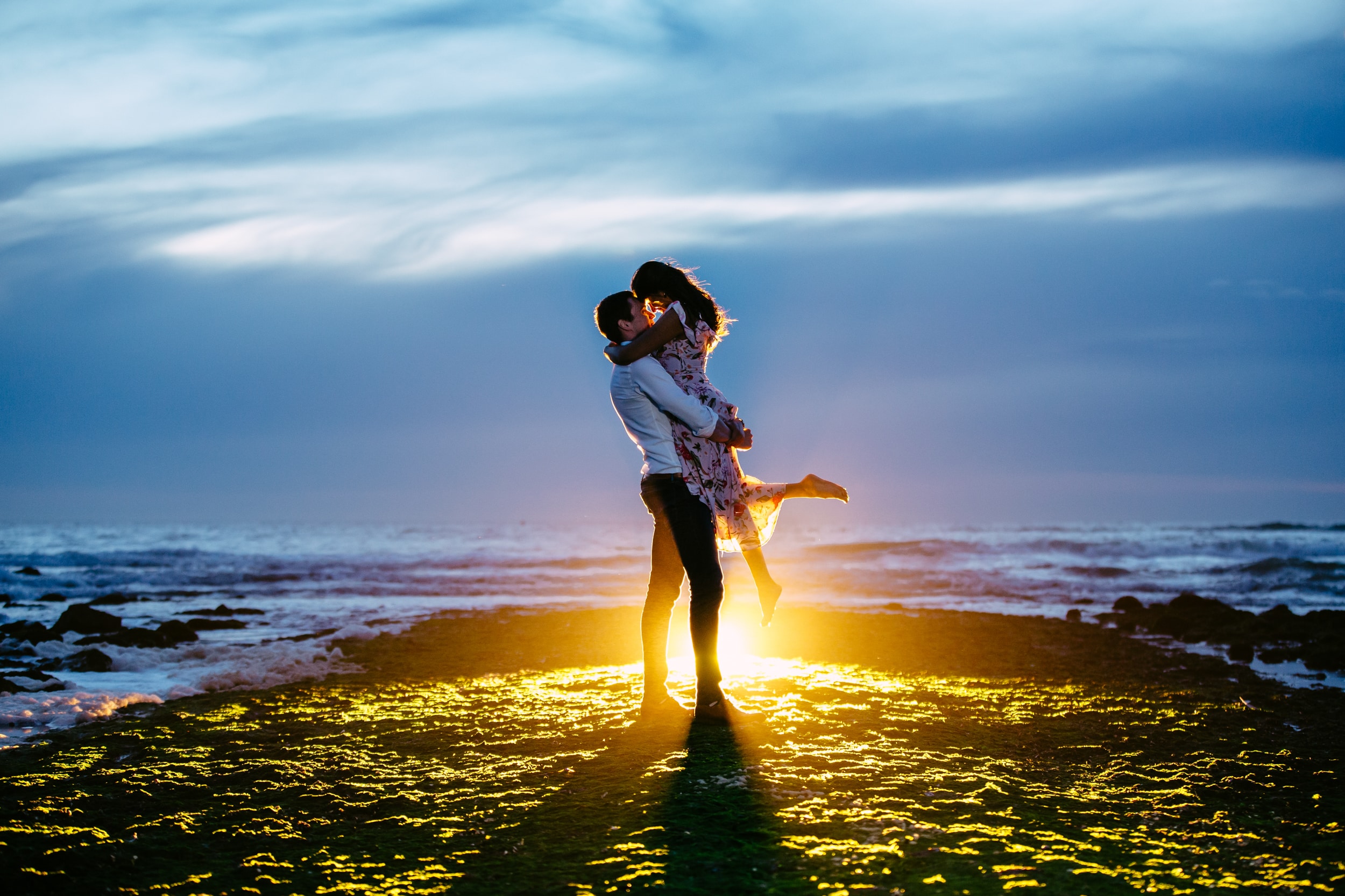 Een man en een vrouw knuffelen elkaar op het strand bij zonsondergang, terwijl hij op het punt staat haar ten huwelijk te vragen.