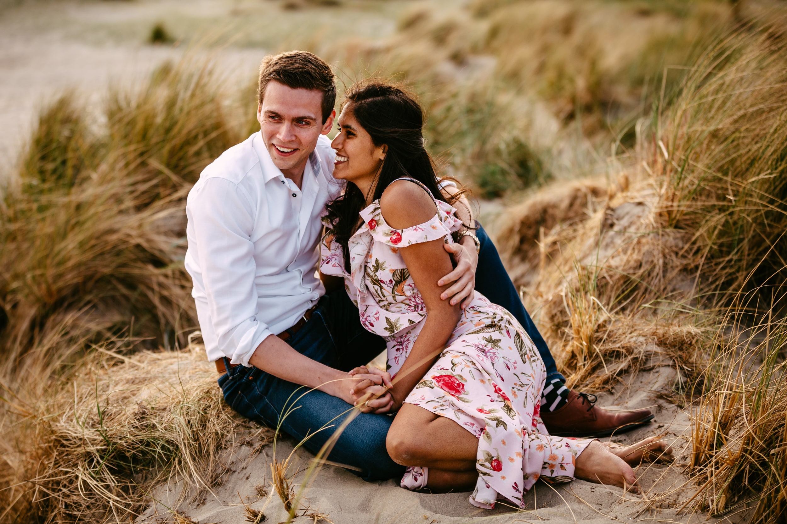 An engaged couple sit in the sand on a beach during their marriage proposal.