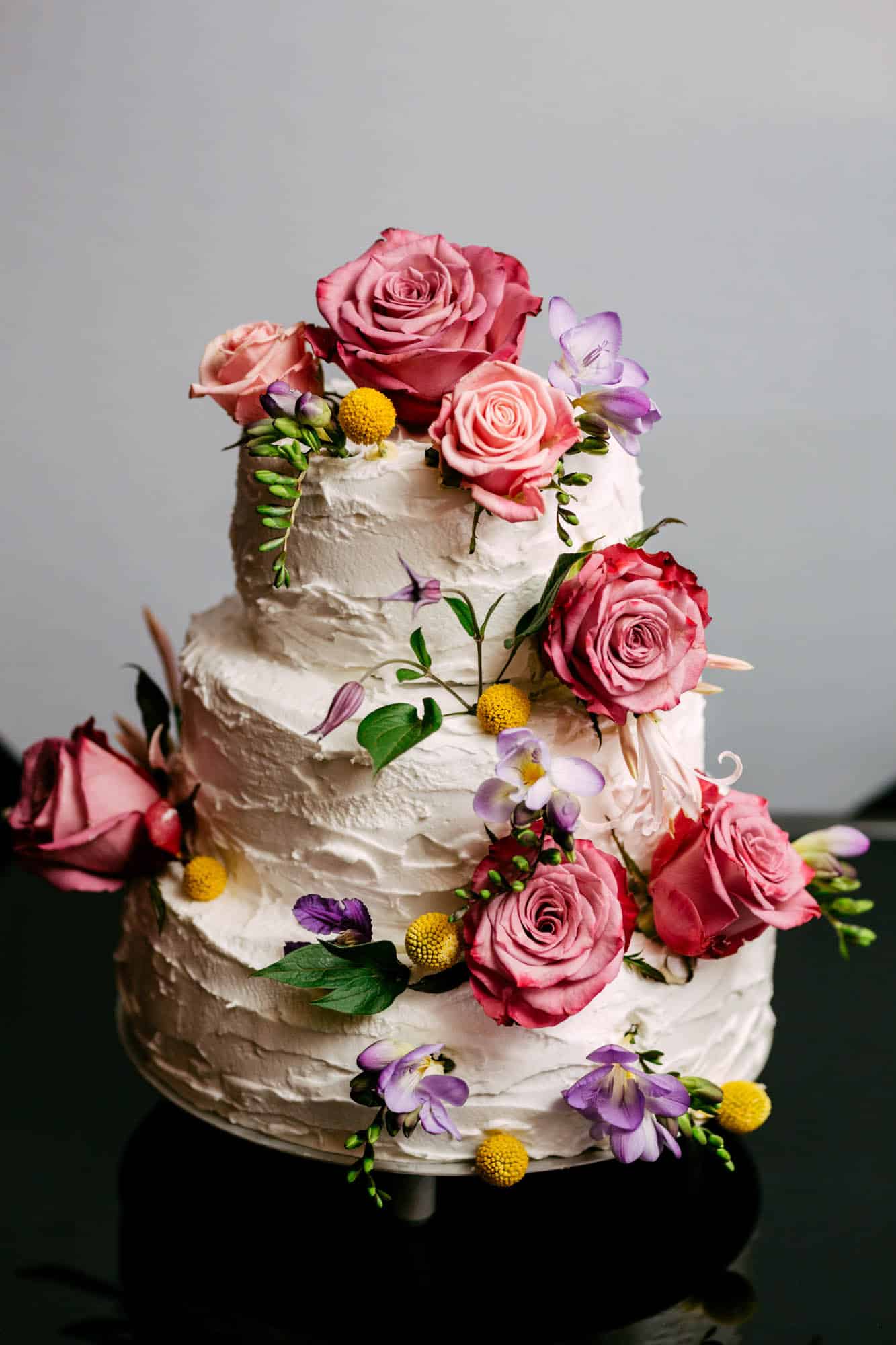 A white wedding cake with pink and purple flowers on top.