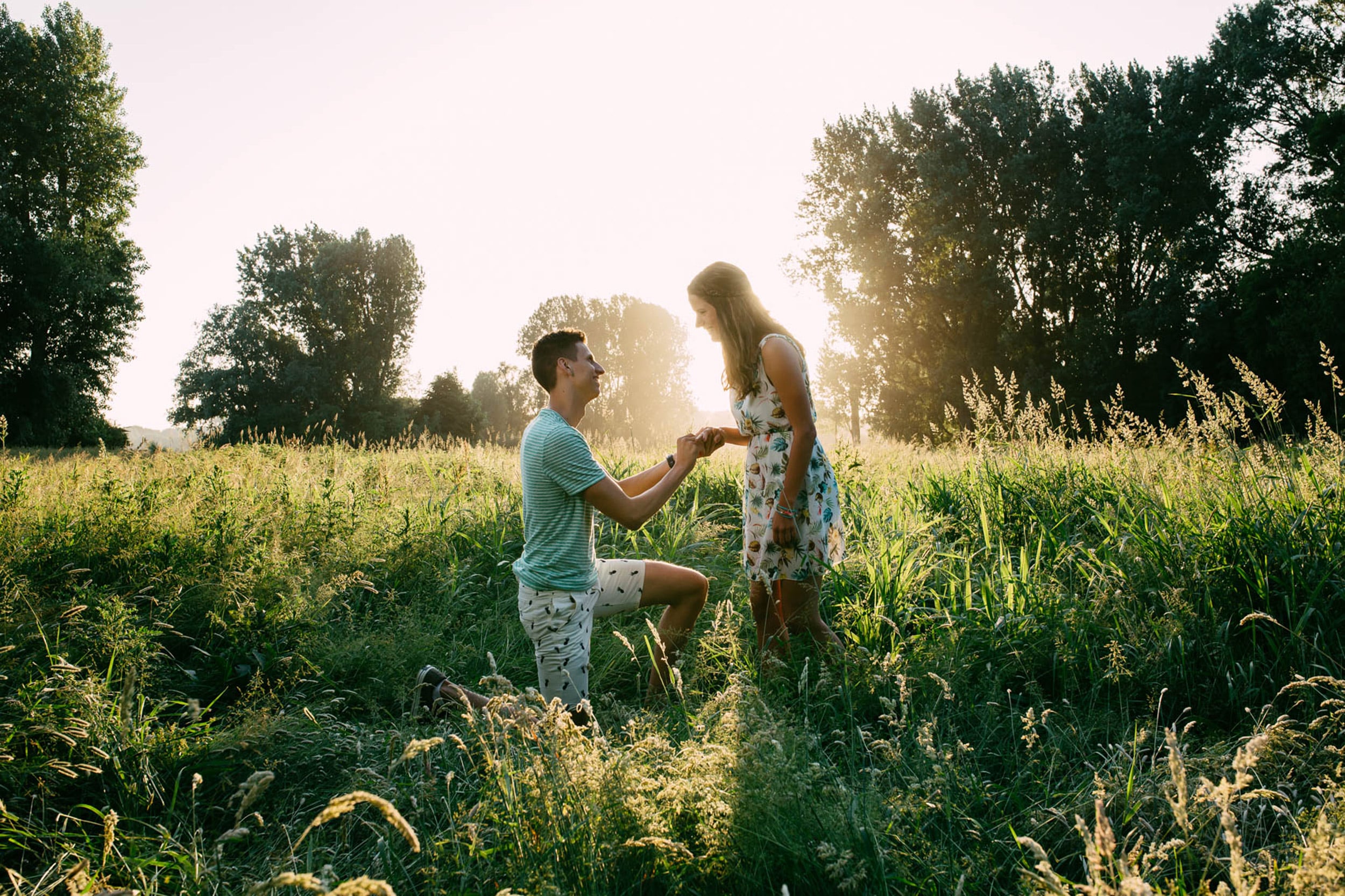A man and a woman kneel down in a field of tall grass and share a blissful moment during their marriage proposal.