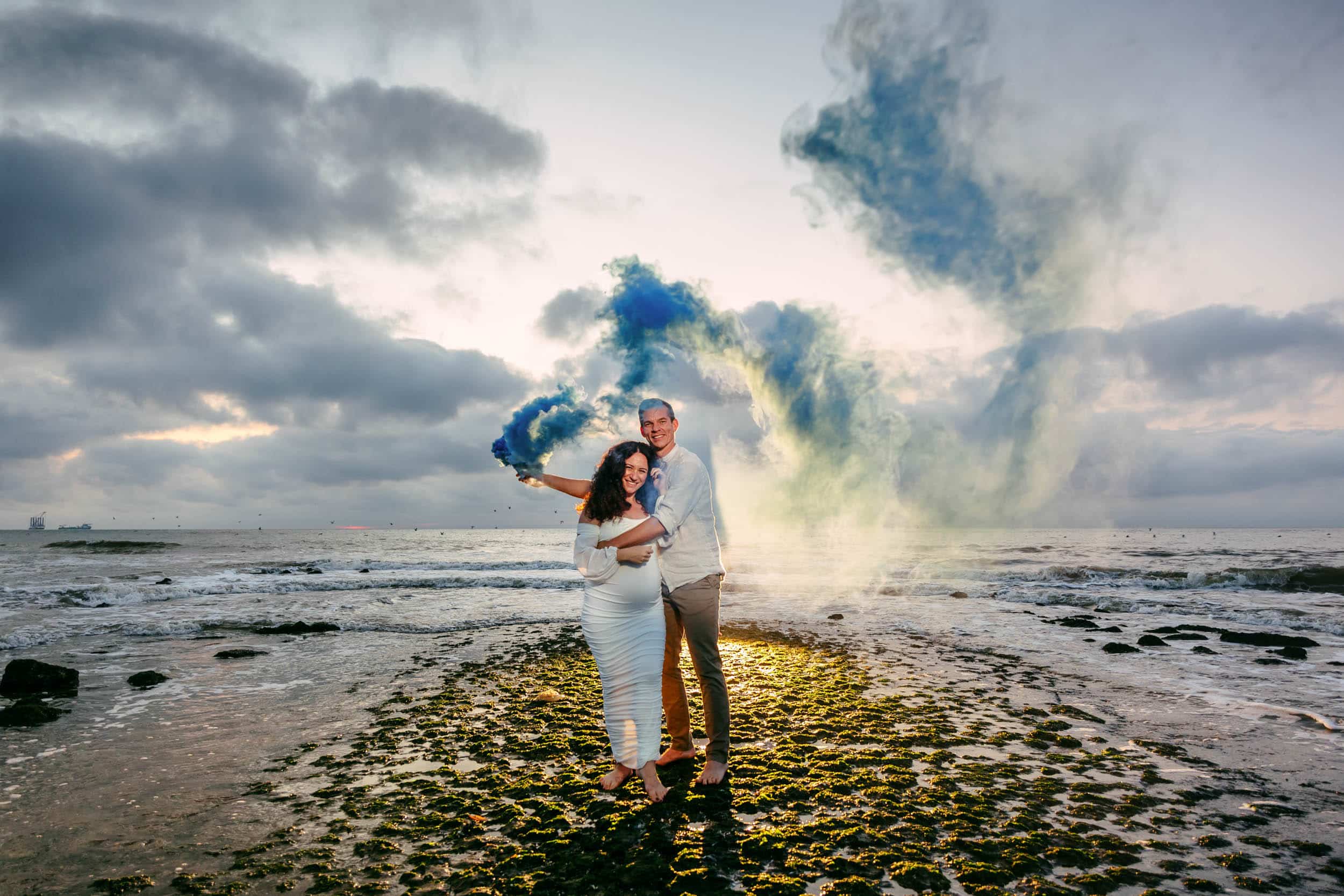 Een stel viert hun zwangerschap met een Zwangerschapsshoot op het strand, vrolijk in hun handen met blauwe rookbommen.