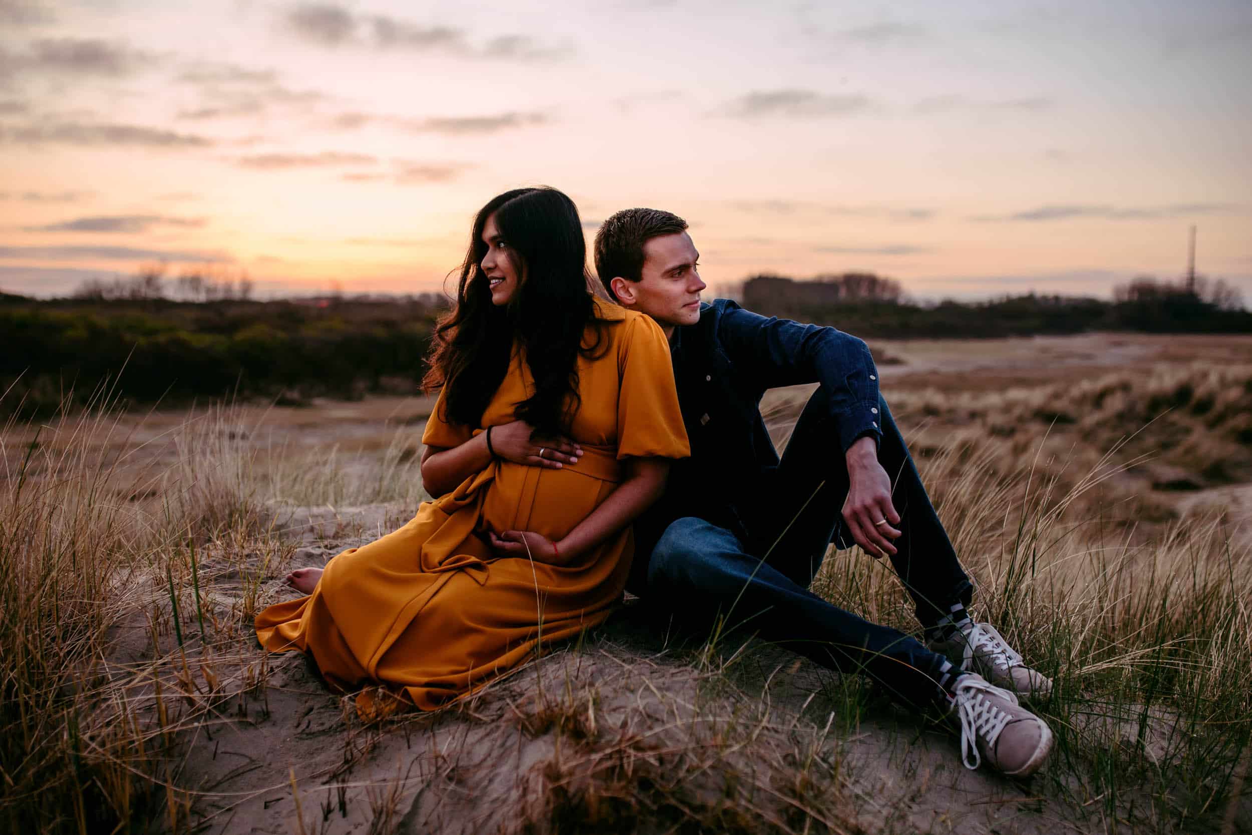 Een zwangerschapsshootpaar zittend in het zand bij zonsondergang.