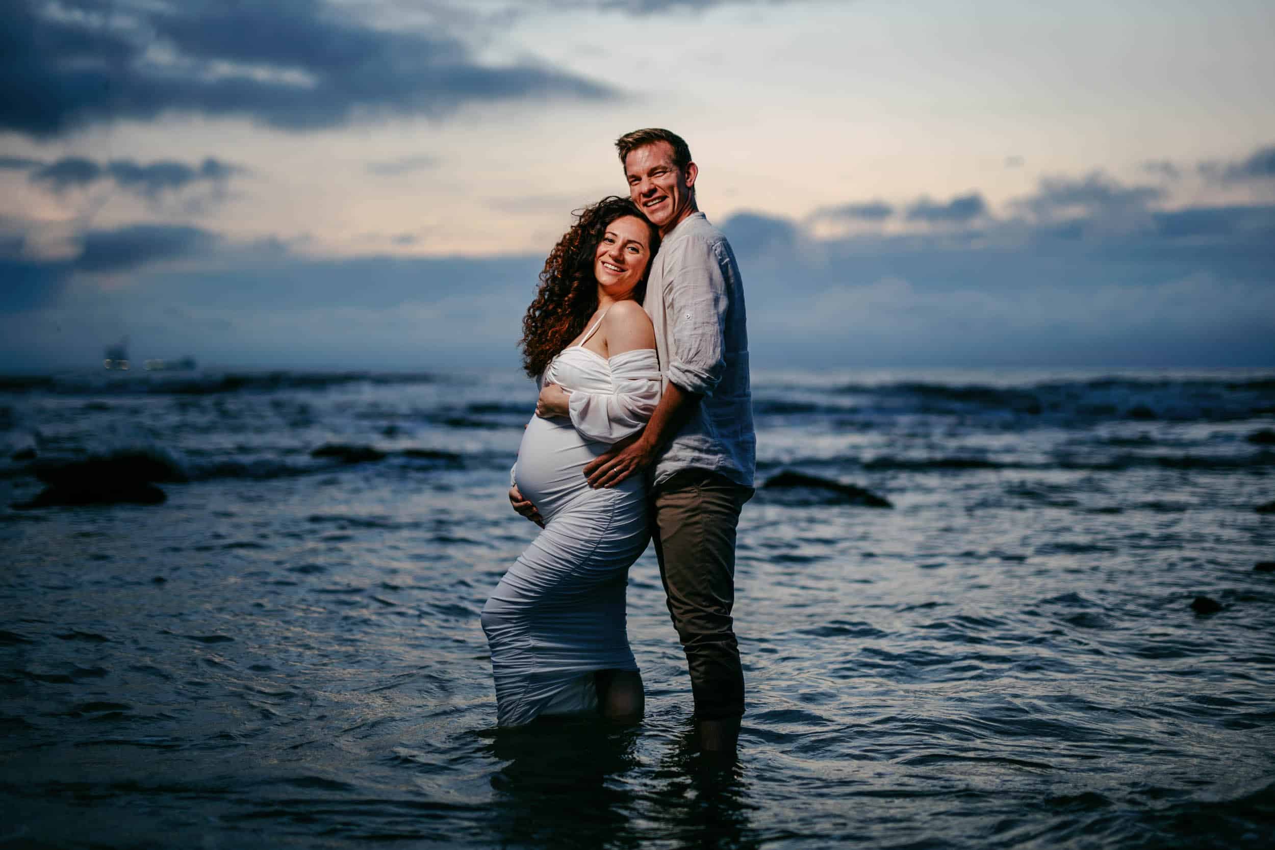 A maternity shoot couple standing in the ocean at sunset.
