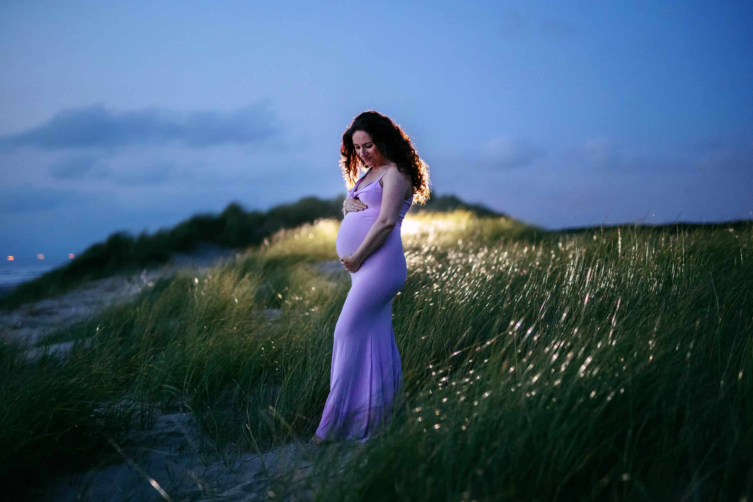 Een zwangere vrouw in een paarse jurk die 's nachts in een veld staat tijdens een Zwangerschapsshoot.