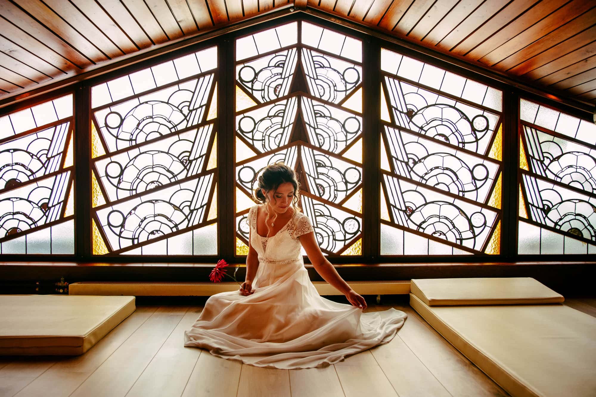 A bohemian bride sits in front of a stained-glass window, dressed in a beautiful wedding dress.