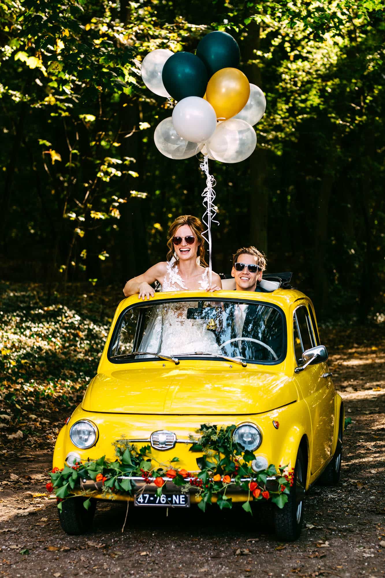 Een bruid en bruidegom in een gele auto met ballonnen vieren hun Trouwen in het bos.