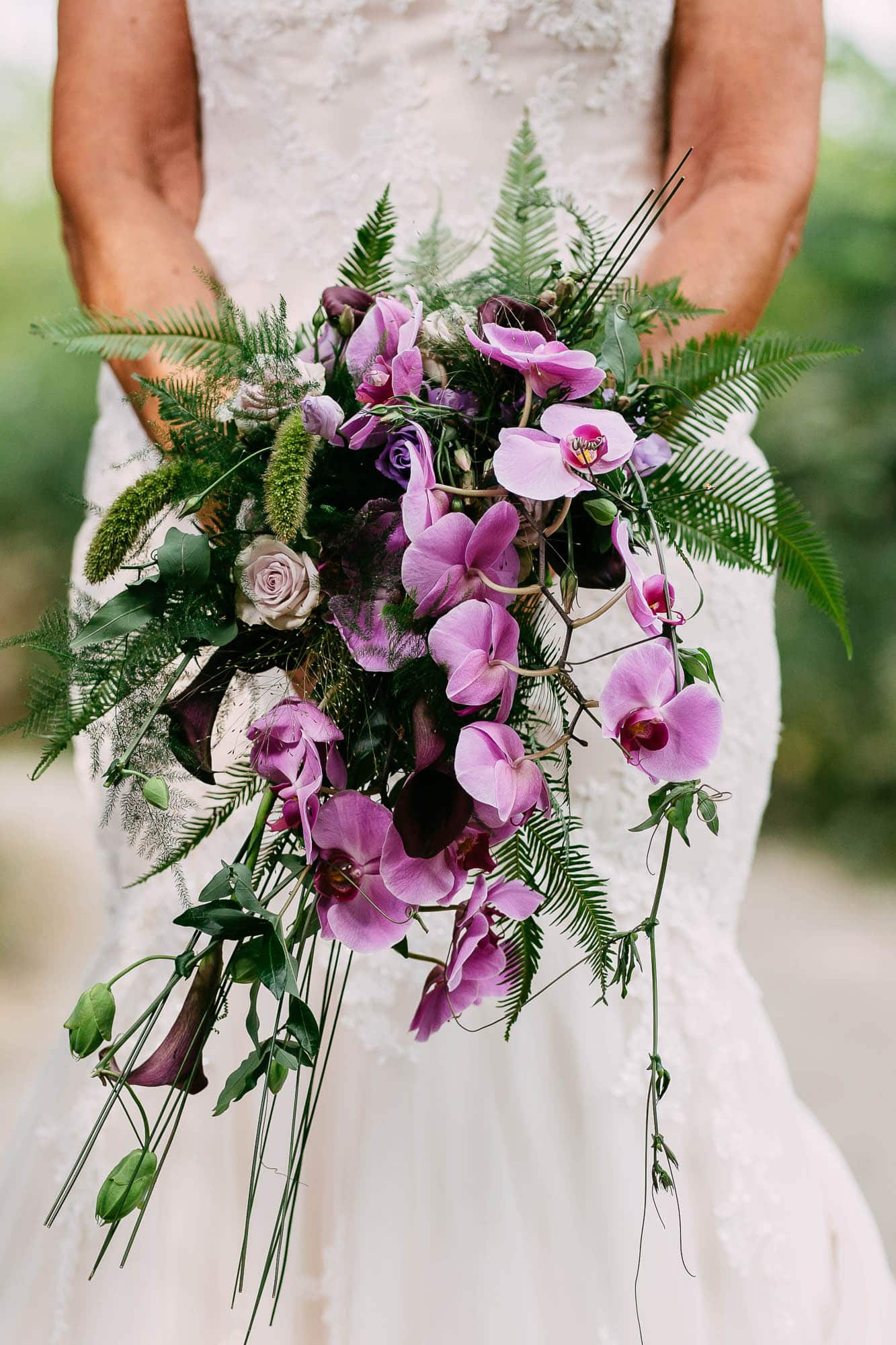 A bride holds a wedding bouquet of purple orchids and ferns.