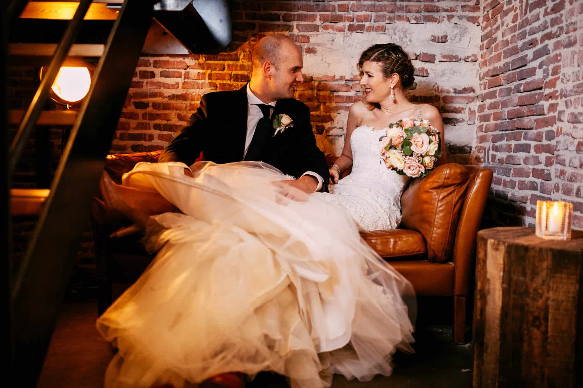 A bride and groom sit happily on a sofa in a beautifully decorated brick room as they plan their wedding.