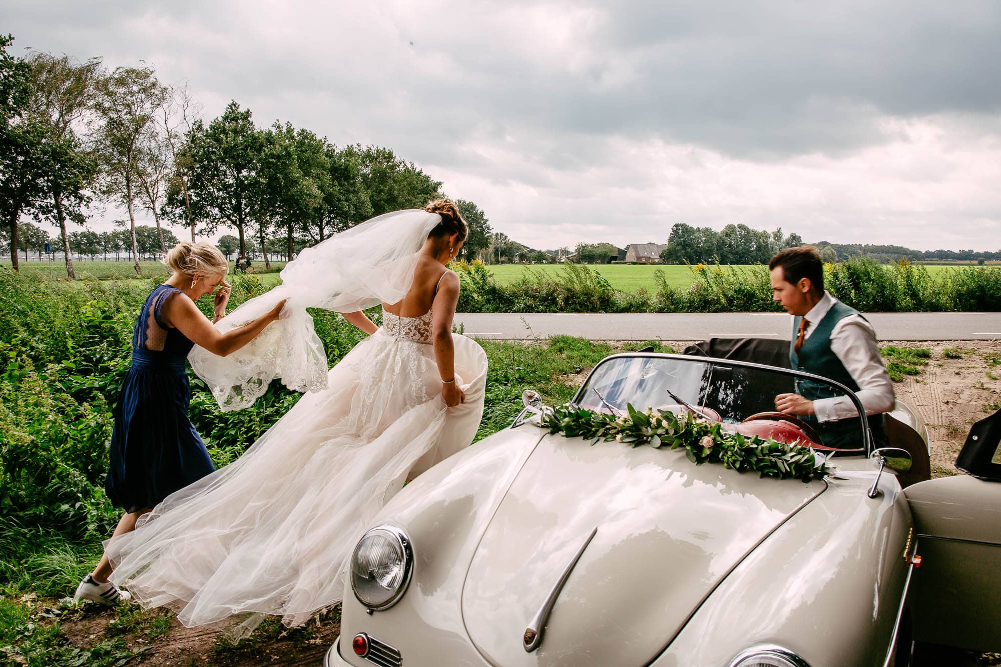 Een bruid en bruidegom maken zich klaar in een oldtimer met hulp van hun ceremoniemeester.