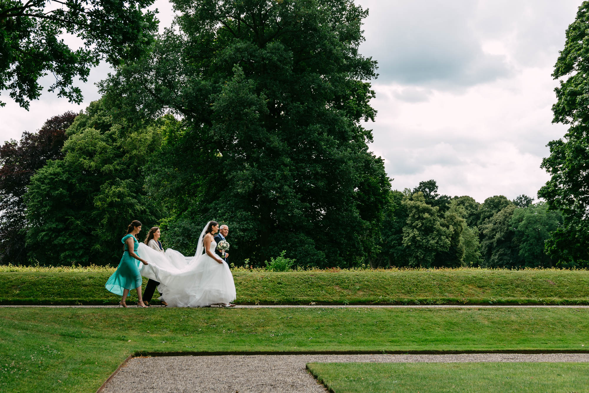 A bride and her master of ceremonies walk through a park.
